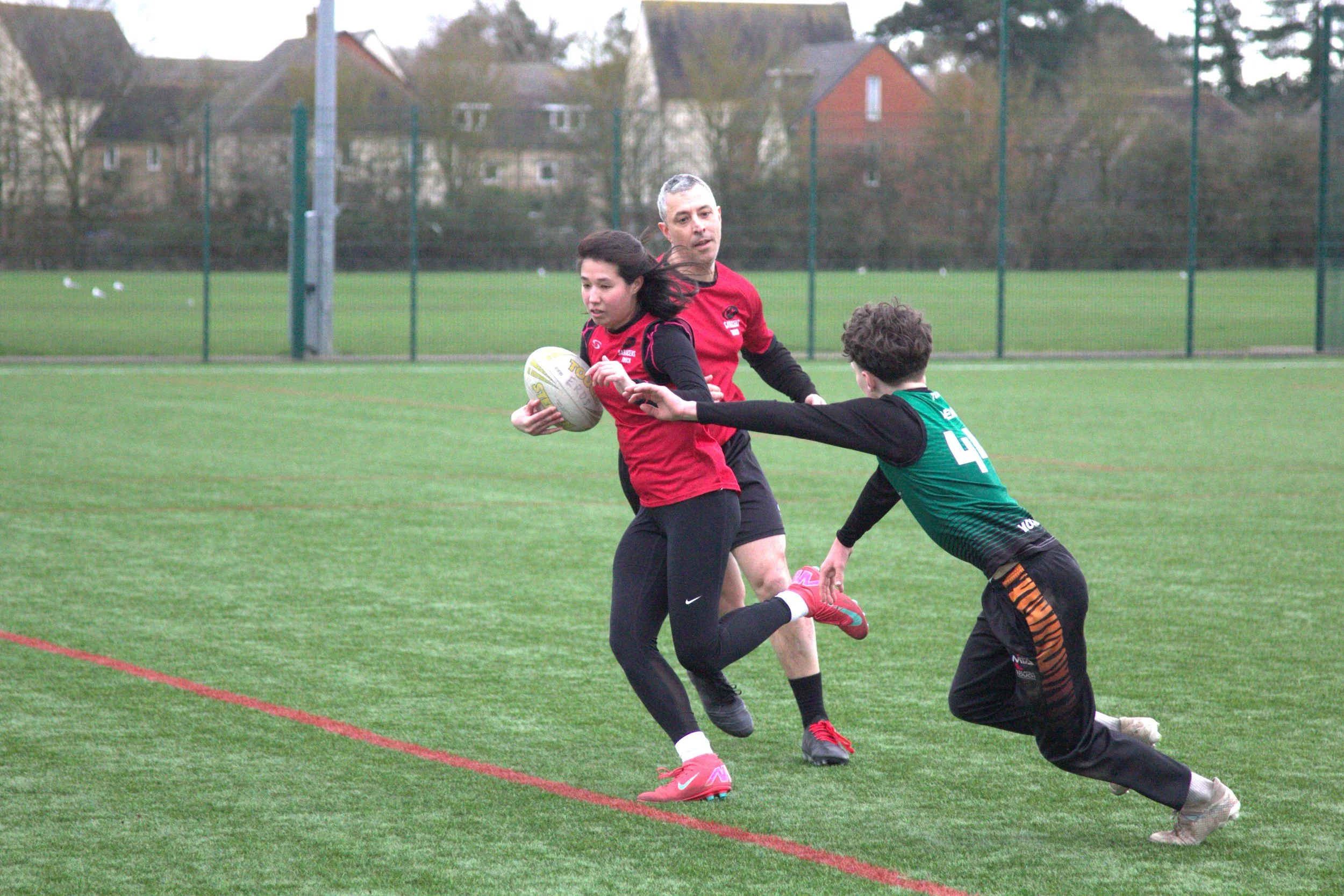 A rugby game with a female player holding a rugby ball, running while being pursued by a male defender, with a coach or referee observing in the background on a grassy field.