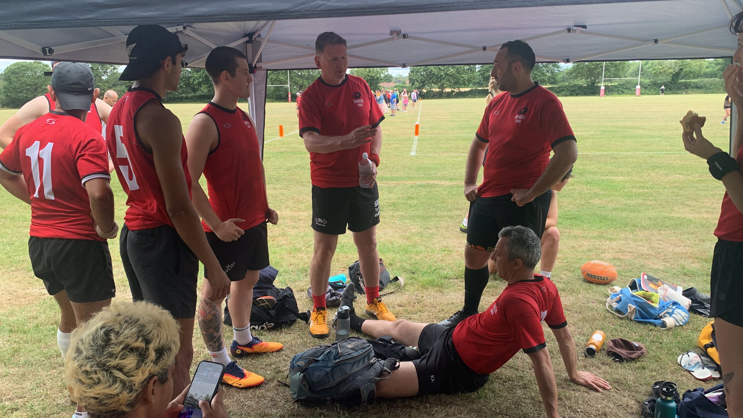 Rugby players in red jerseys and black shorts gathered under a tent on a grassy field during a break in play, with some sitting and others standing, surrounded by water bottles, bags, and personal items, on a cloudy day.