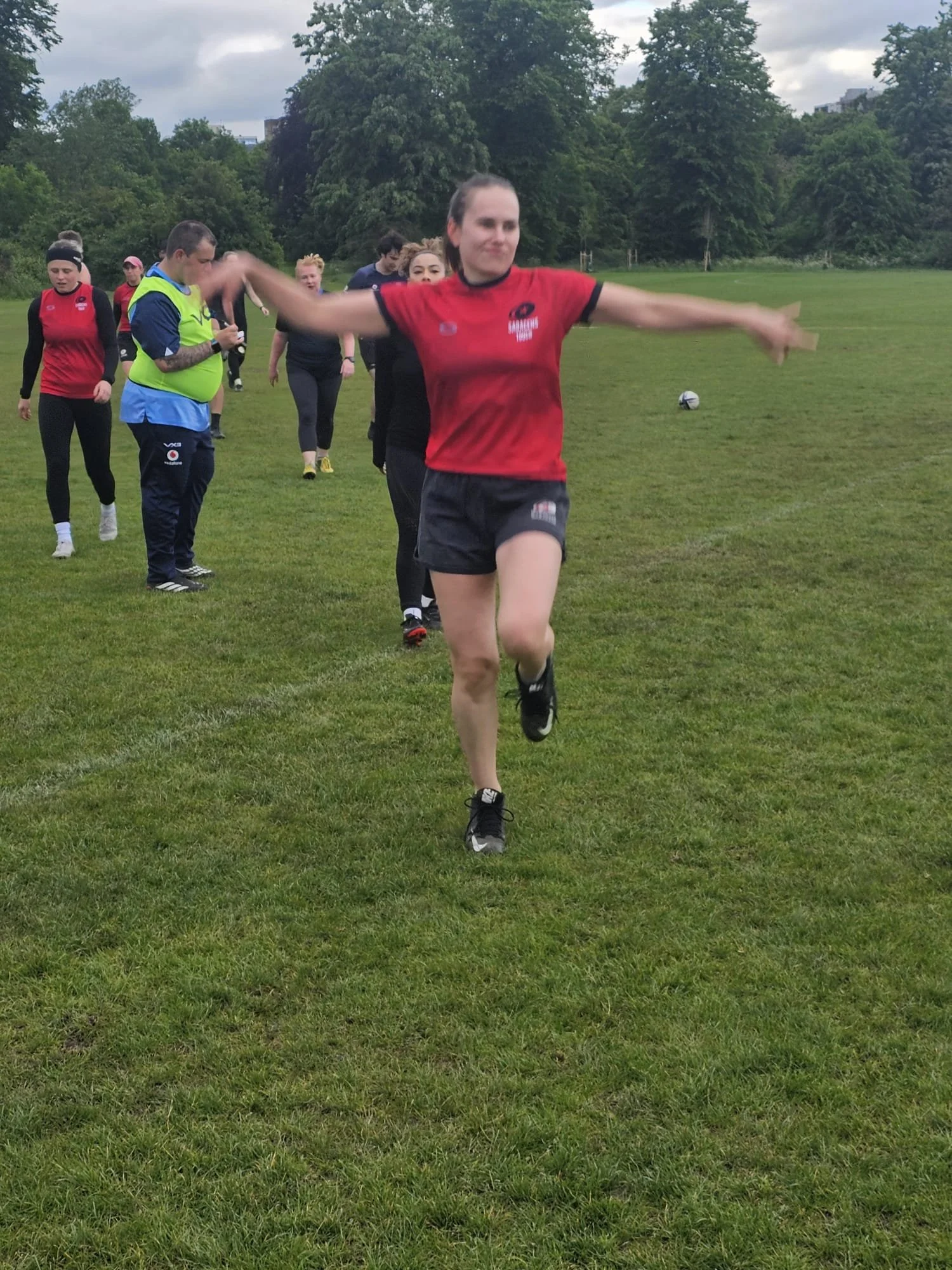A group of women participating in an outdoor rugby practice or warm-up on a grassy field, with one woman in a red shirt and black shorts running toward the camera.