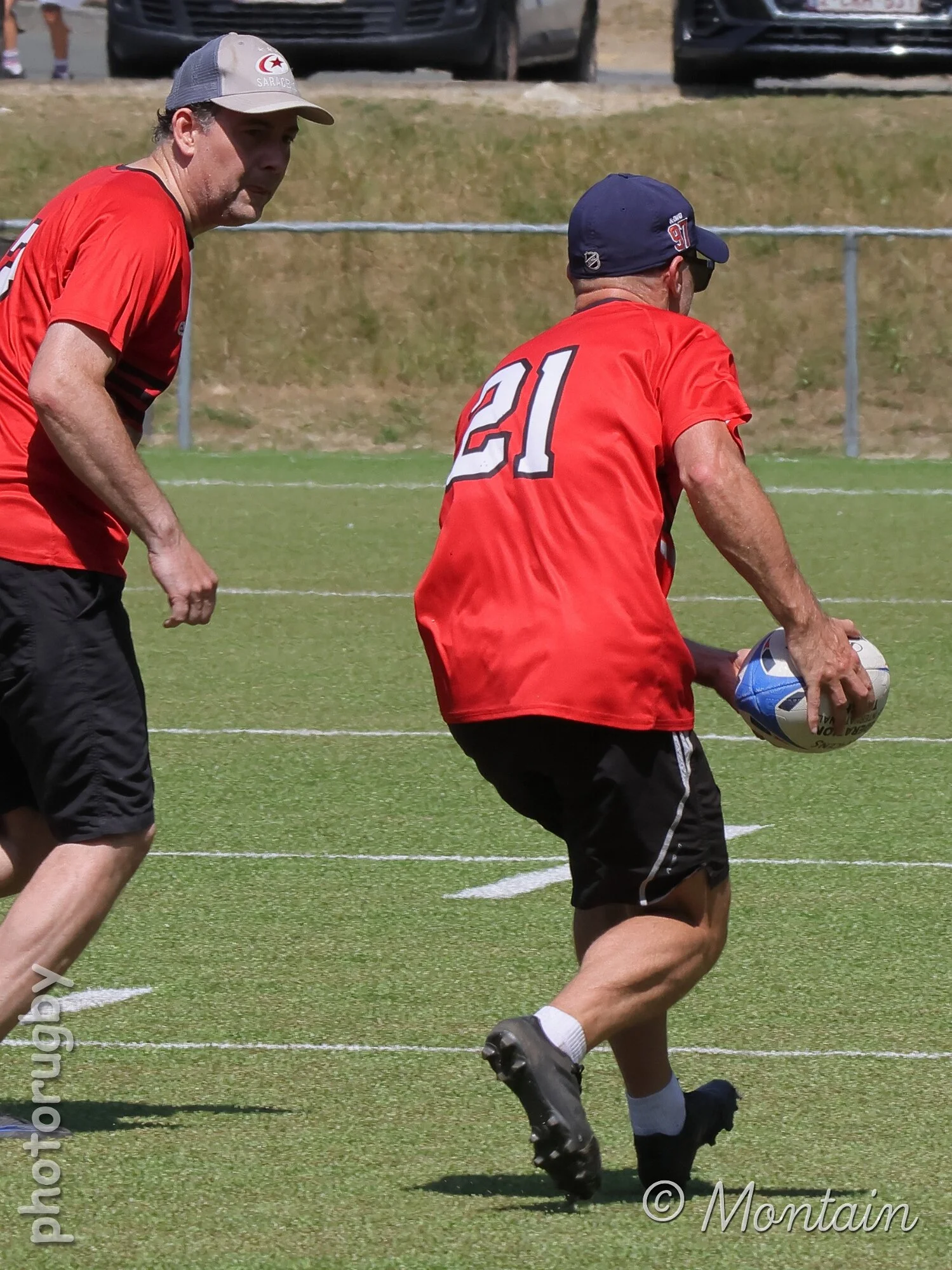 Two men playing rugby on a grass field. One man is wearing a red shirt with the number 21, black shorts, and a navy cap, holding a rugby ball. The other man is wearing a red shirt, black shorts, and a trucker cap, standing nearby.
