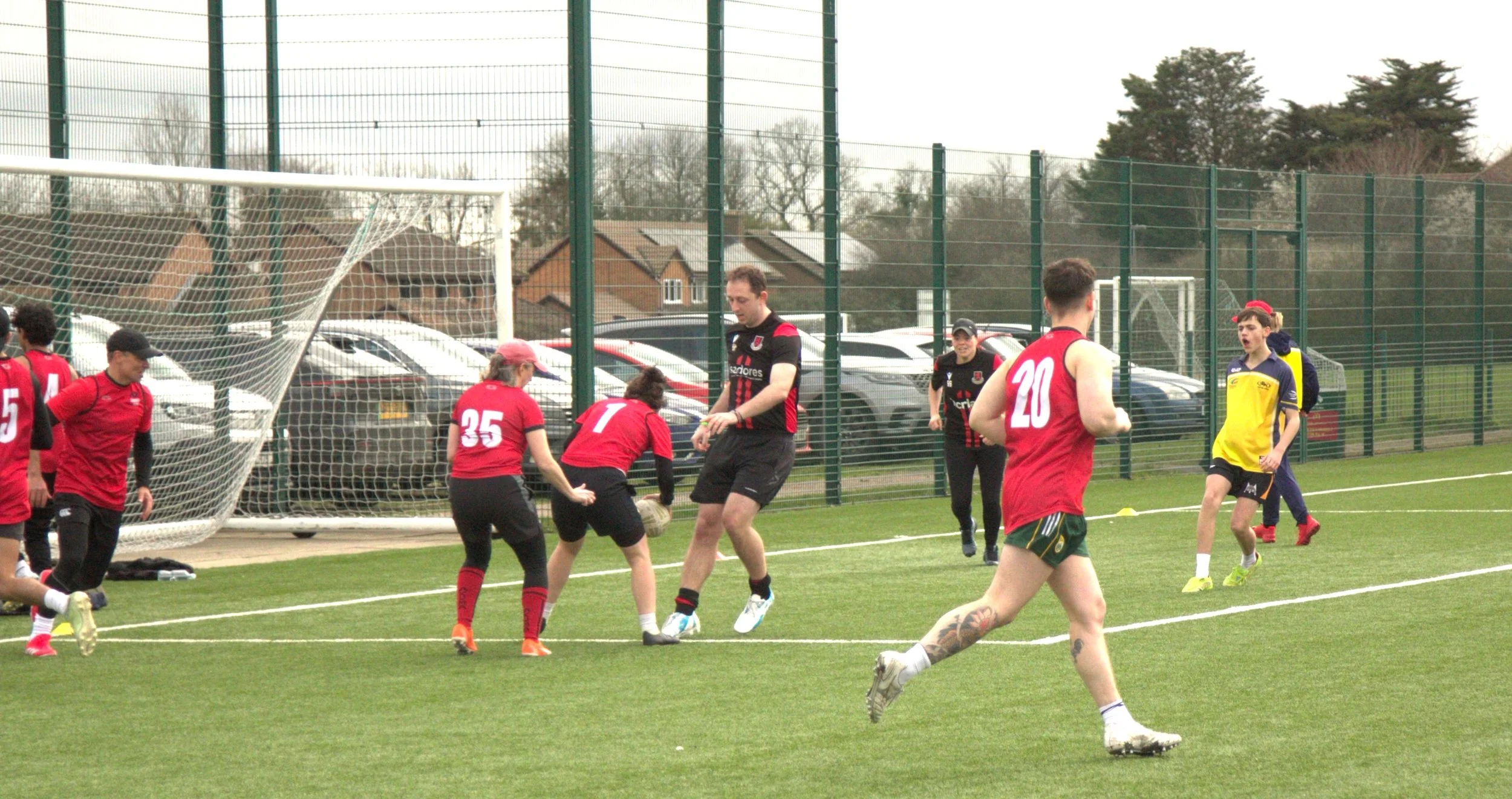 People playing cricket on a sports field, some wearing red jerseys and some in yellow and blue, with a goal post and parking lot in the background.