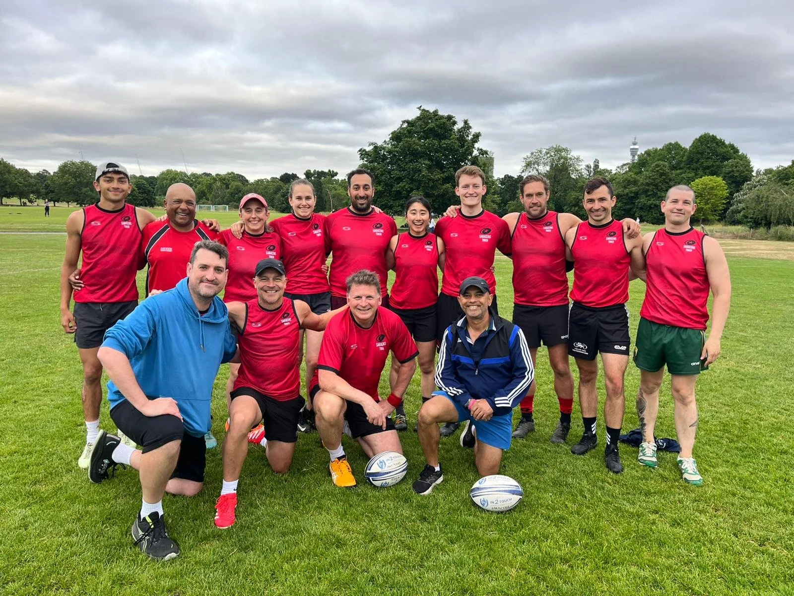 A group of people in sportswear standing on a grassy field, posing for a photo, with cloudy skies and trees in the background.