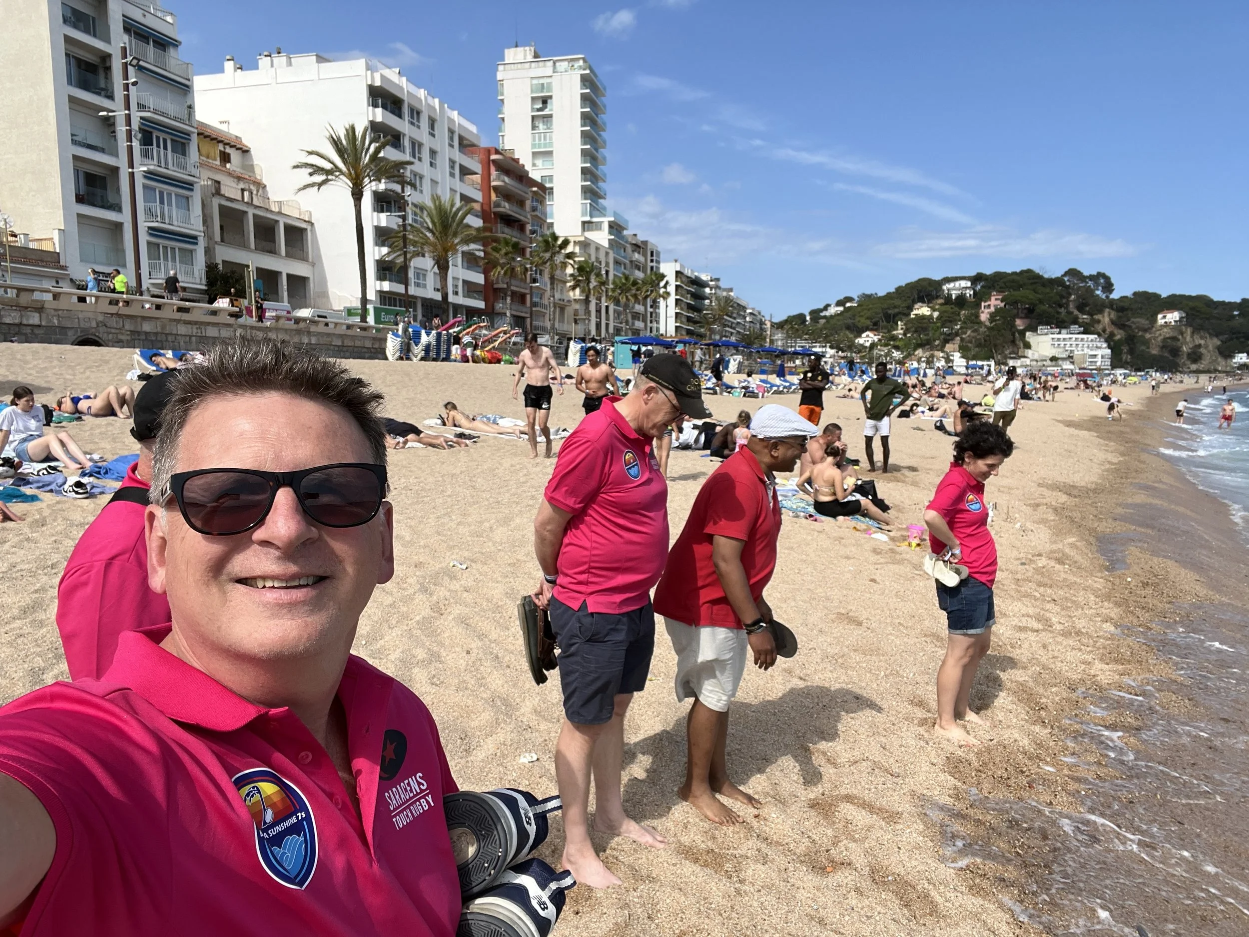 A man taking a selfie on a crowded beach with others standing near the shoreline, some in pink shirts, and high-rise buildings and palm trees in the background.