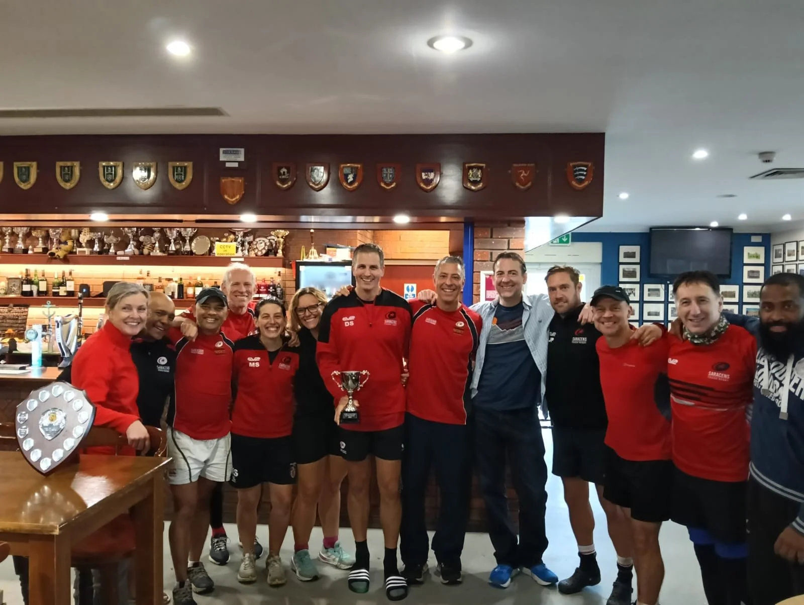Group of people standing together inside a bar or pub, many wearing sports jerseys, some holding a trophy, smiling for a photo.