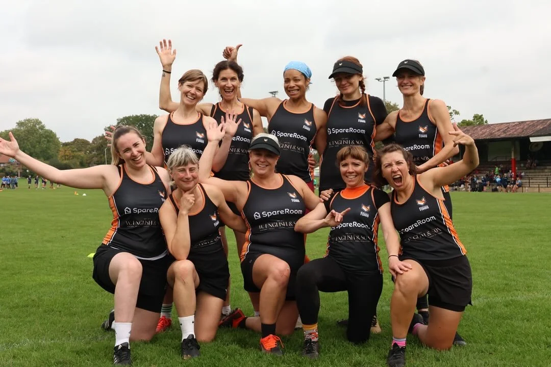 A group of nine women in athletic uniforms celebrating on a sports field, some kneeling and others standing, smiling and making celebratory gestures.