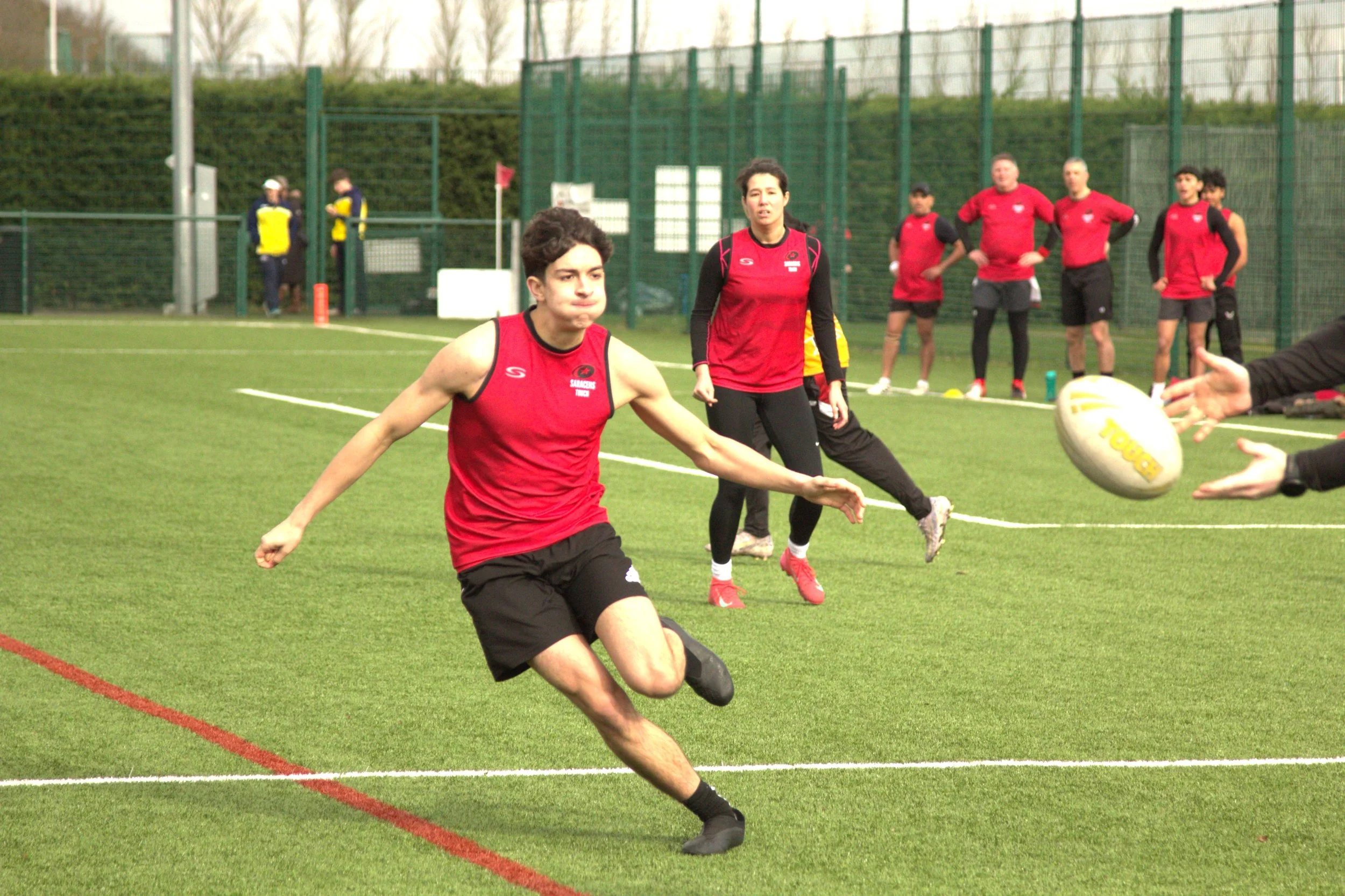 Rugby players in red jerseys participating in a game on a green field, with a focus on a young male player kicking the ball.