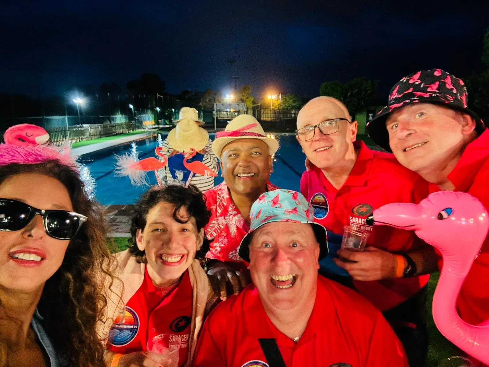 Group of people at a pool party dressed in red shirts and summer accessories, smiling with inflatable flamingos, including a pink flamingo float, at night with pool and fenced area in the background.