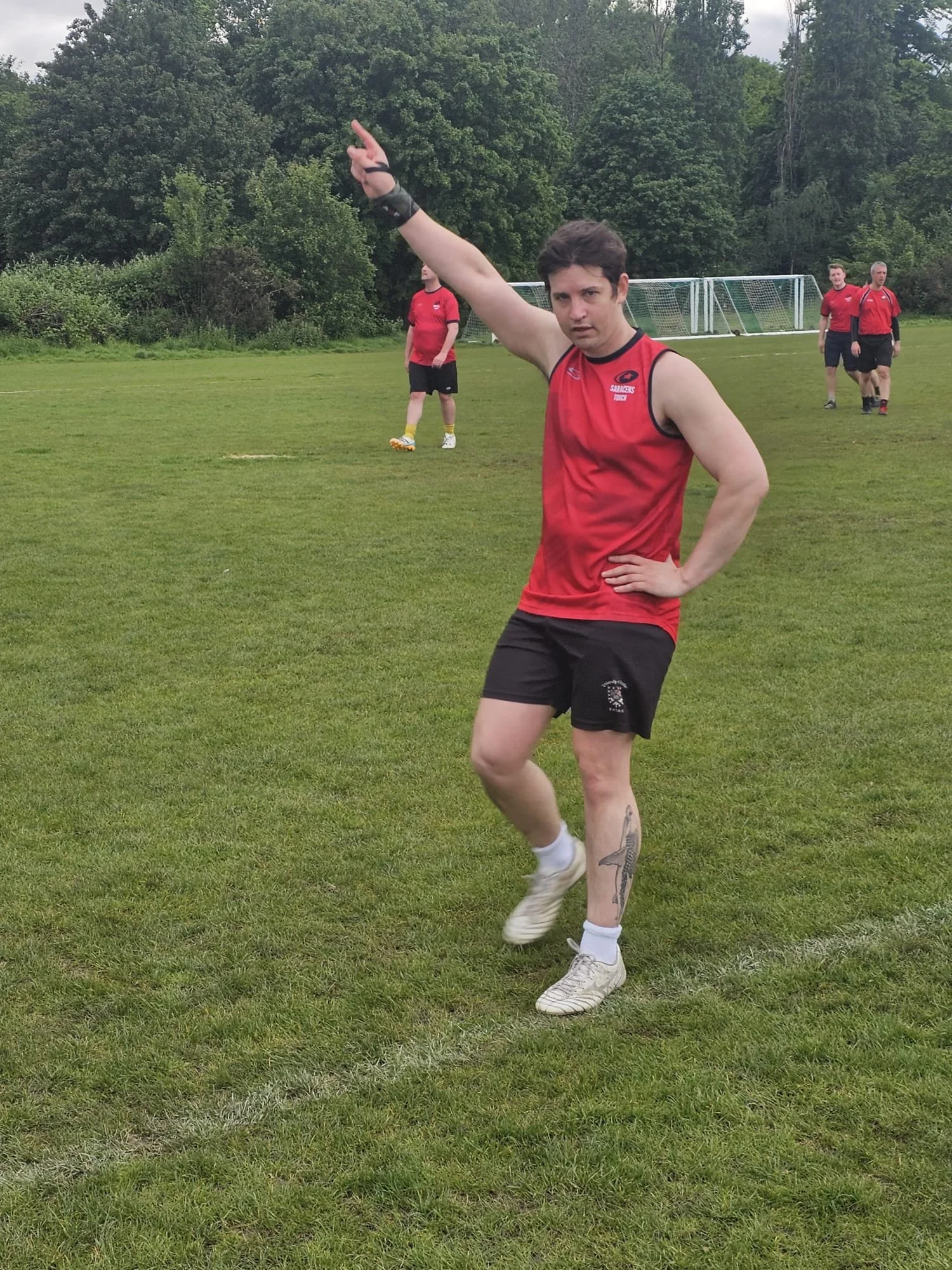 A man in a red athletic jersey and black shorts standing on a grass field, making a gesture with his left arm raised and pointing up, with three other men in similar attire in the background.