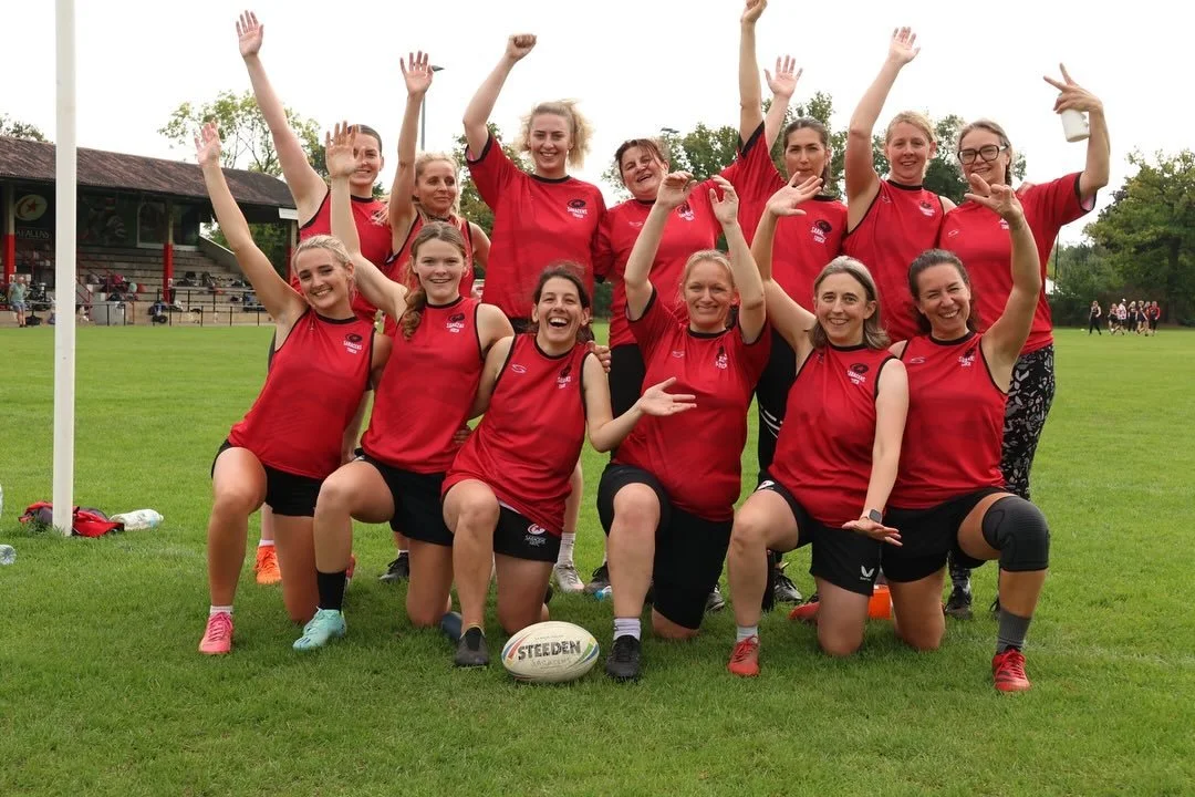 A group of women rugby players in red jerseys celebrating on a grass field with a rugby ball, some kneeling and others standing, raising their arms and smiling.