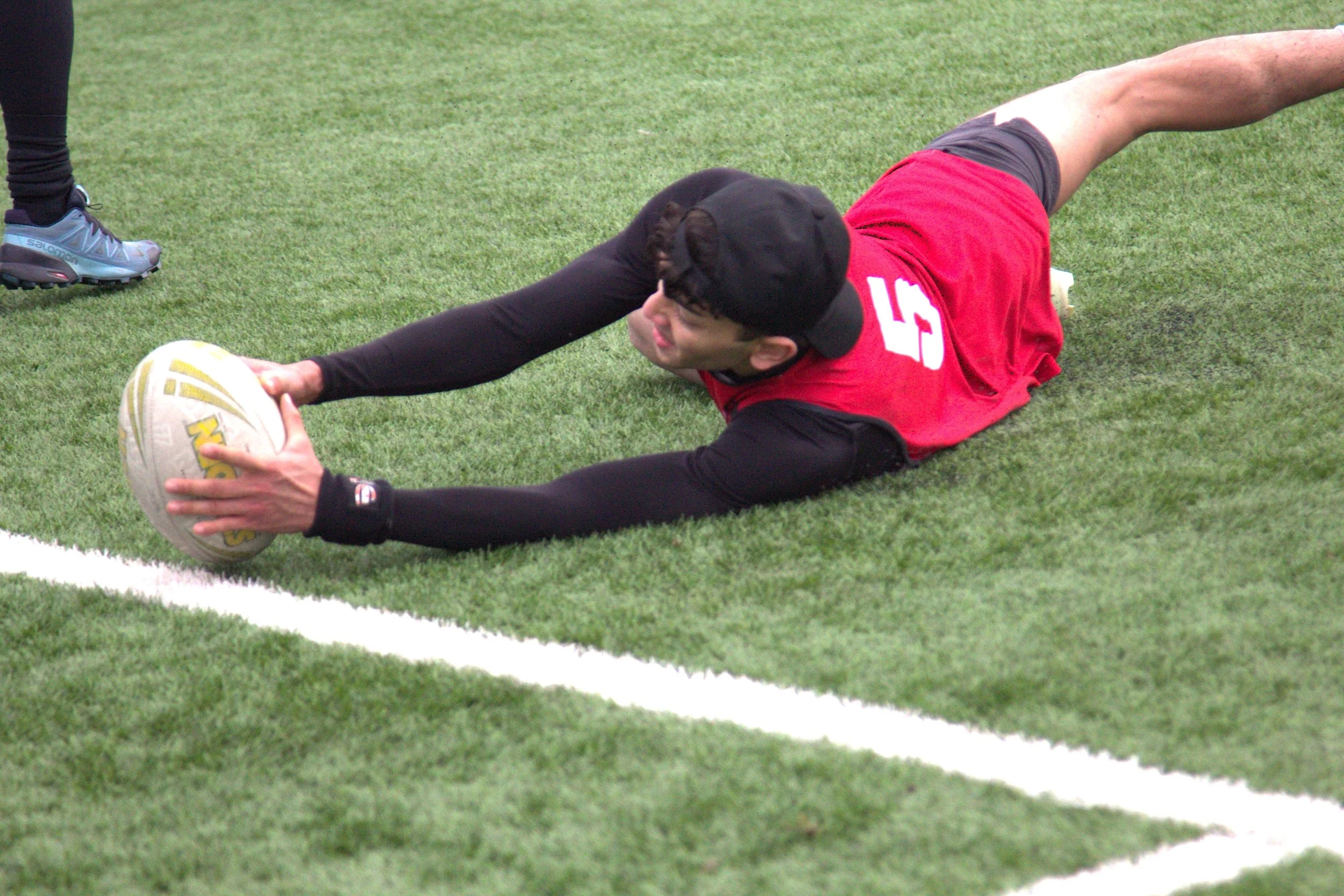 A person wearing a red sports jersey, black cap, and black long sleeve shirt is diving on the field, reaching out with both hands to catch a rugby ball. The person is on the grass in an athletic stance, fingers on the ball, with another individual pa