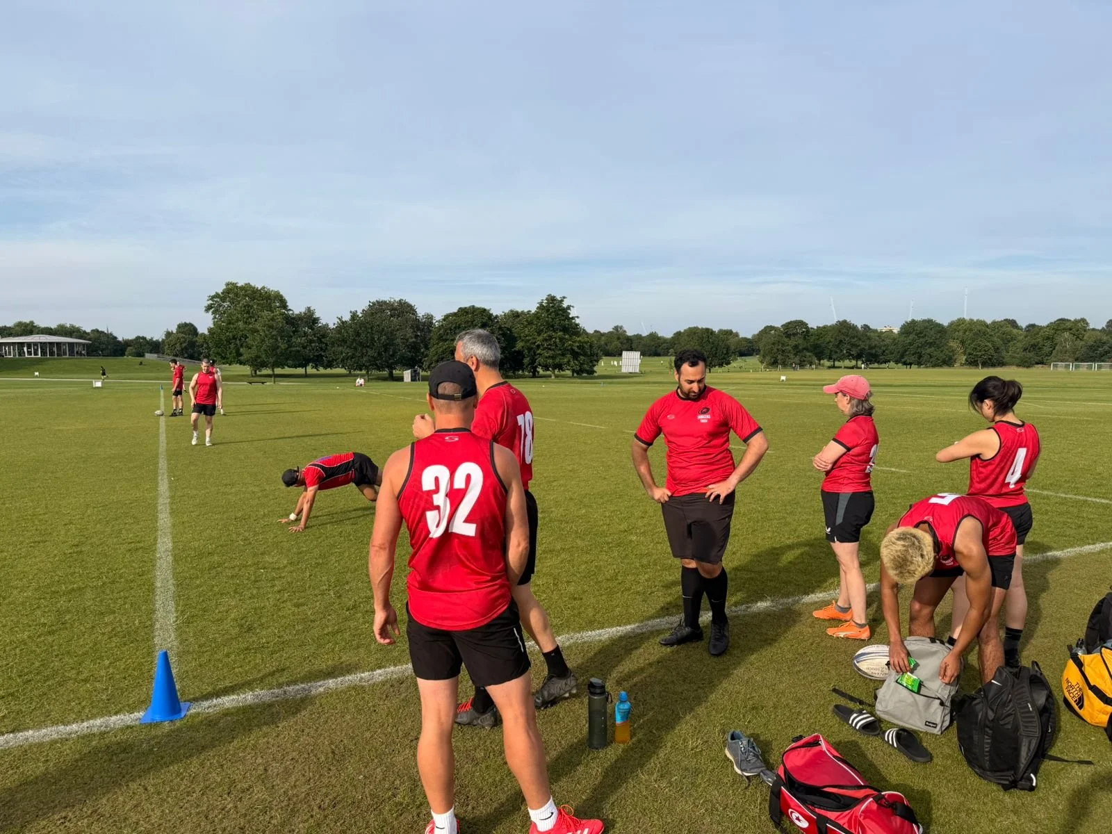 A group of rugby players wearing red jerseys standing on a green field with trees in the background, some stretching, drinking water, and preparing for the game.