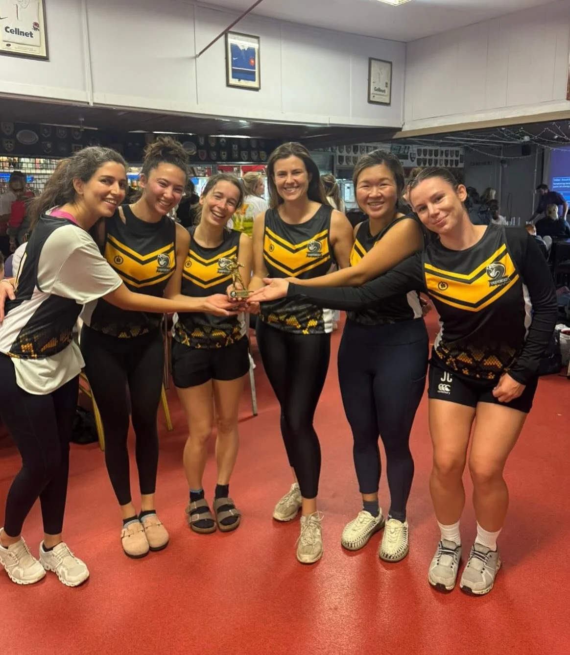 Group of six women in black and yellow sports uniforms holding a small trophy together in a sports venue.