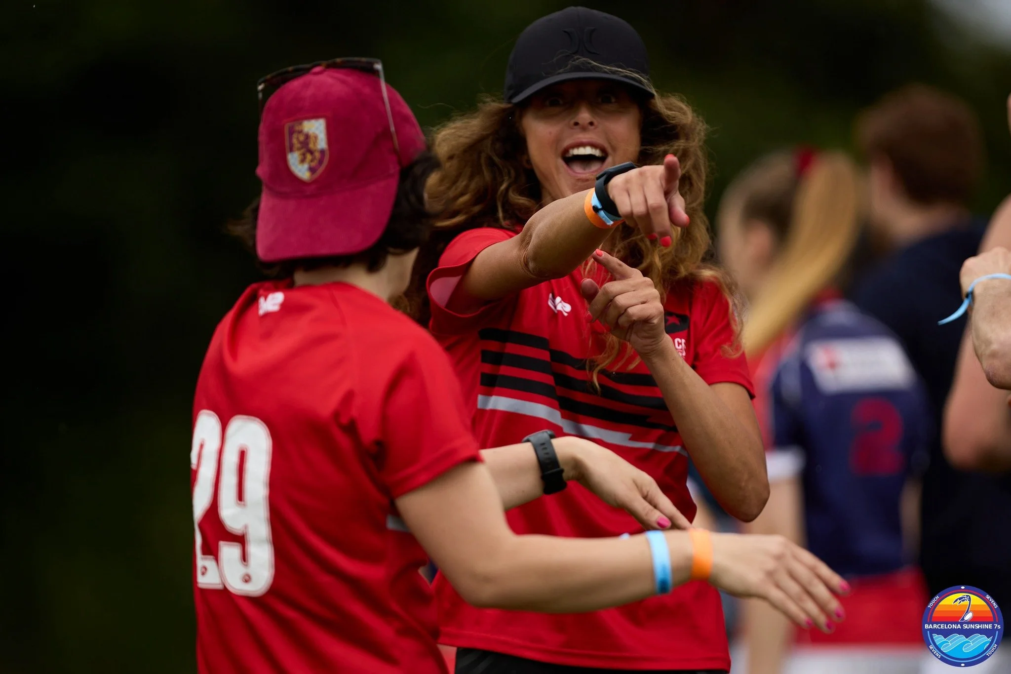 A woman with curly hair and a black cap, wearing a red sports shirt, is pointing and speaking to a young girl with a red cap and shirt, during a sports event. Several other women are visible in the background.