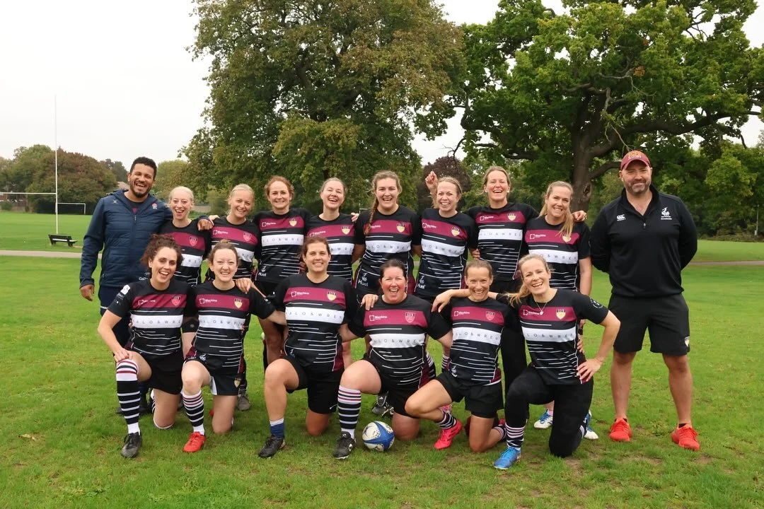 A women's rugby team with two coaches standing and kneeling on a grassy field, smiling and posing for a group photo, surrounded by trees.
