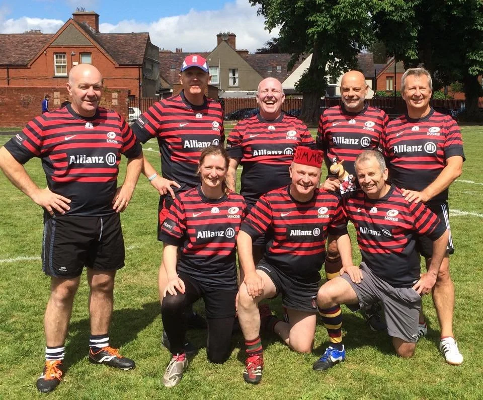 Group of eight adults in black and red striped rugby jerseys on a grassy field, smiling, with houses and a tree in the background.