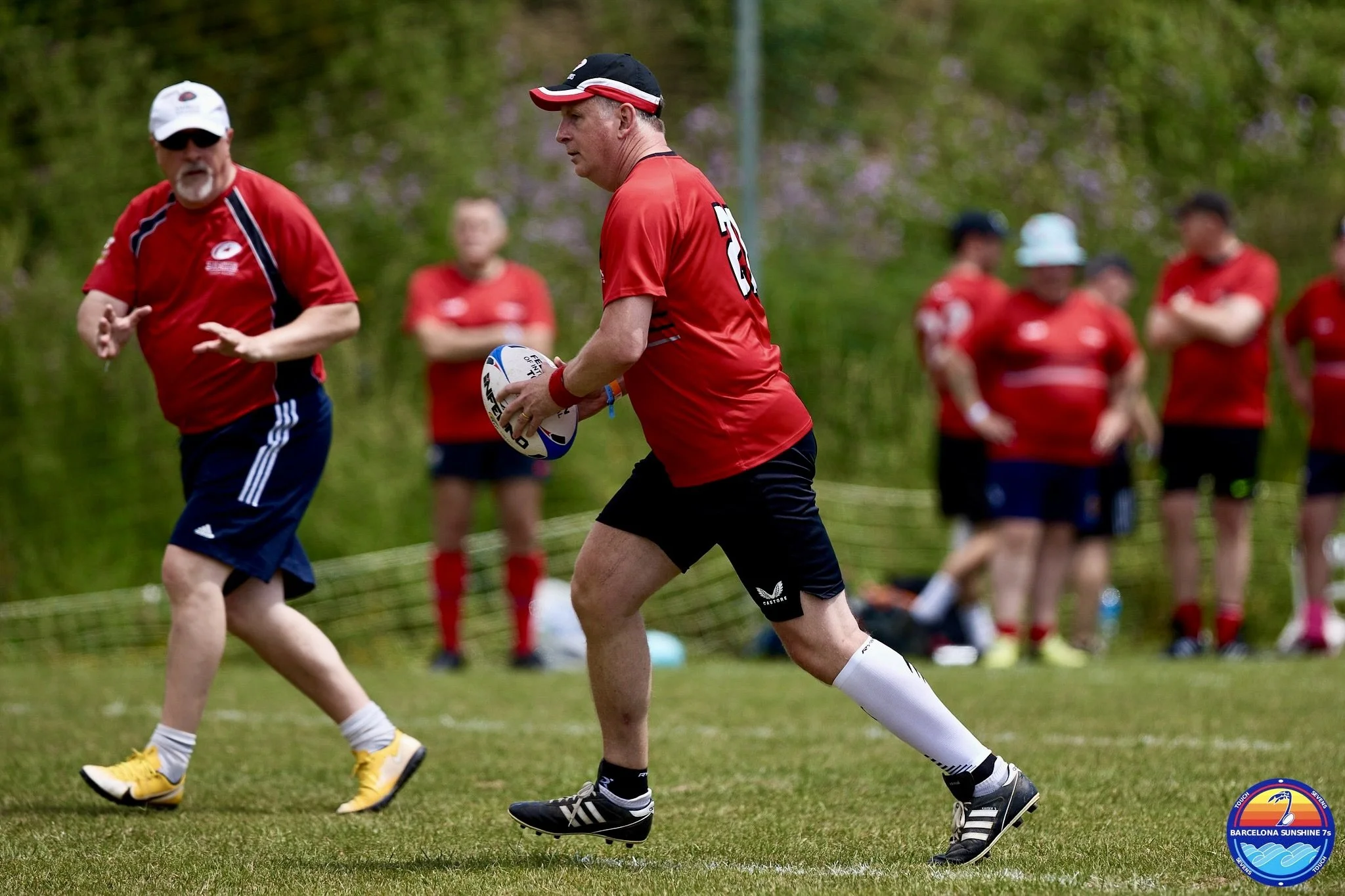 A man in red sports uniform holding a rugby ball on a grassy field during a game, with other players and spectators in the background.