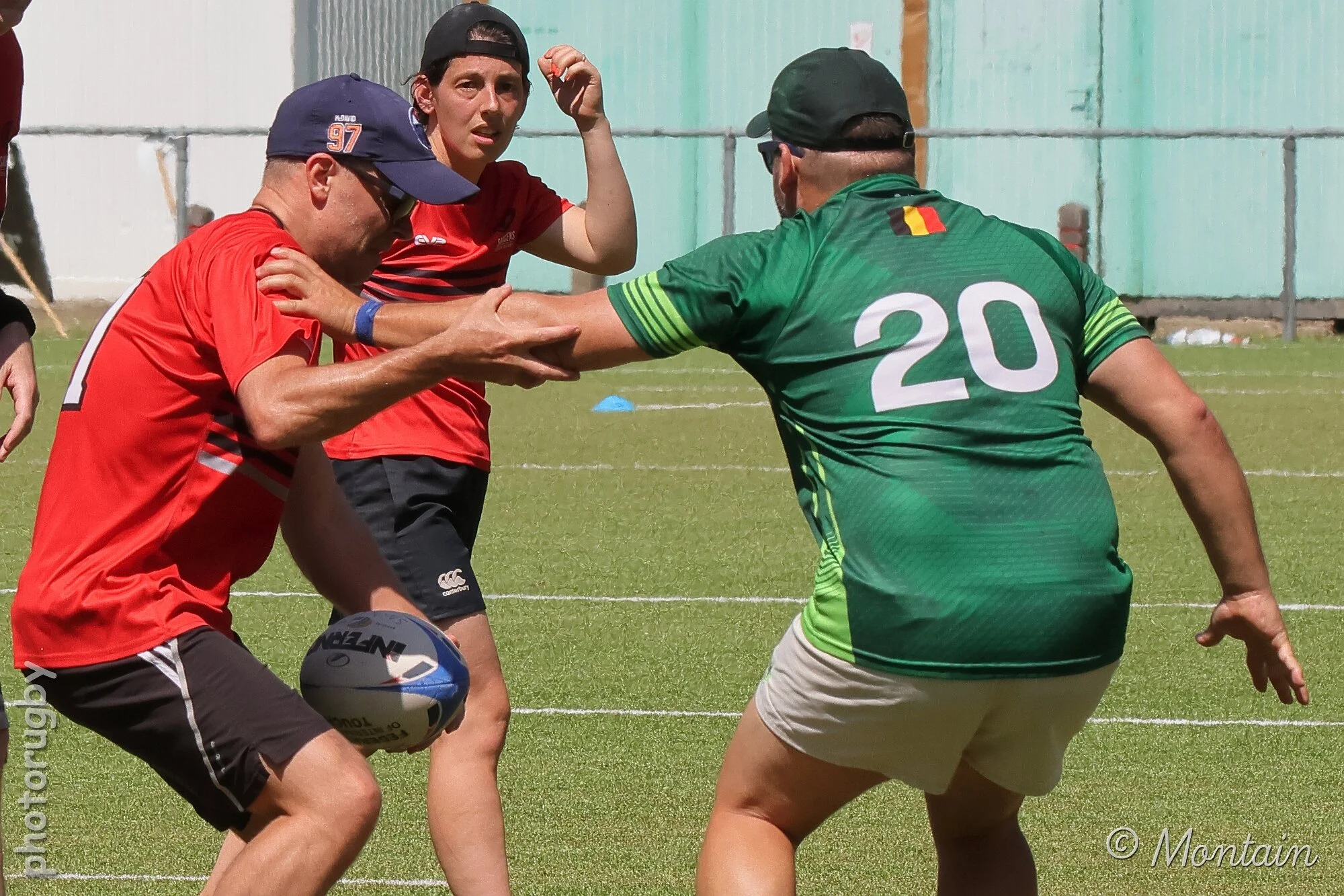 Three people playing rugby on a field, one with a football, two attempting to tackle or block the person with the ball. All are wearing sports clothing and caps.