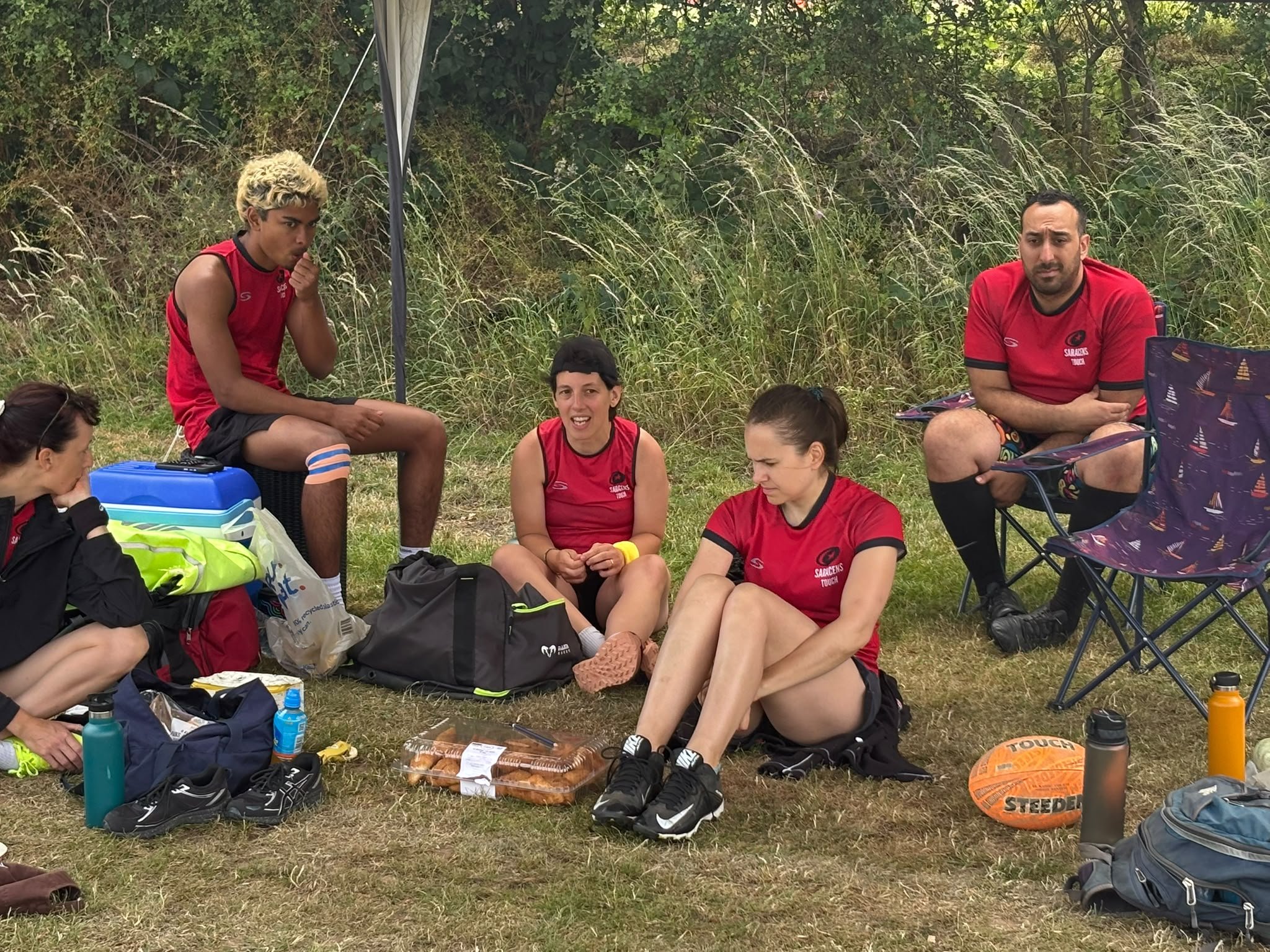 A group of young people sitting and standing on grass outdoors, wearing red sports uniforms with black accents, amidst sports equipment and bags, with trees and tall grass in the background.