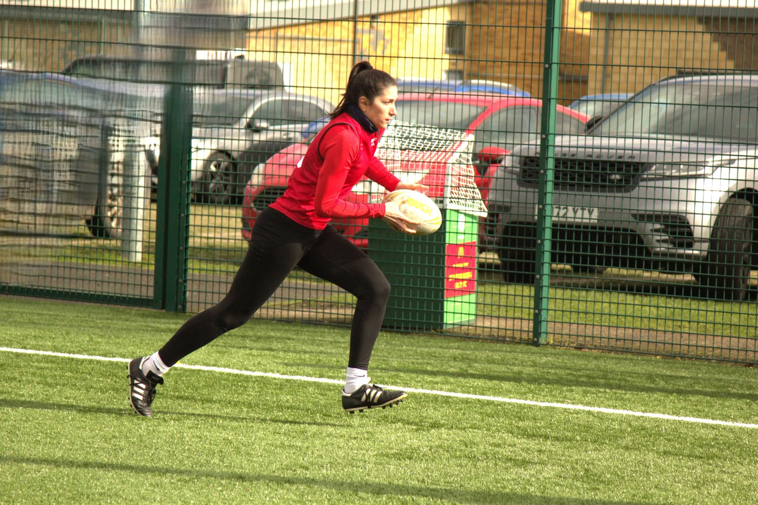 A woman in a red jacket and black pants playing rugby, running with a rugby ball on a field with a fence and parked cars in the background.