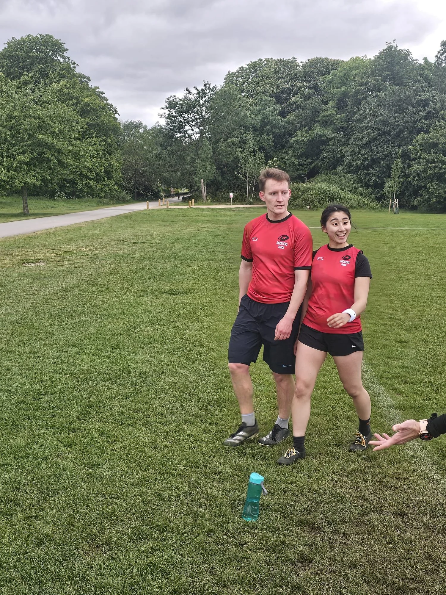 Two young people in athletic clothing standing in a park with green grass and trees, under cloudy skies. One is a man with light skin and short hair, the other is a woman with darker hair, smiling.