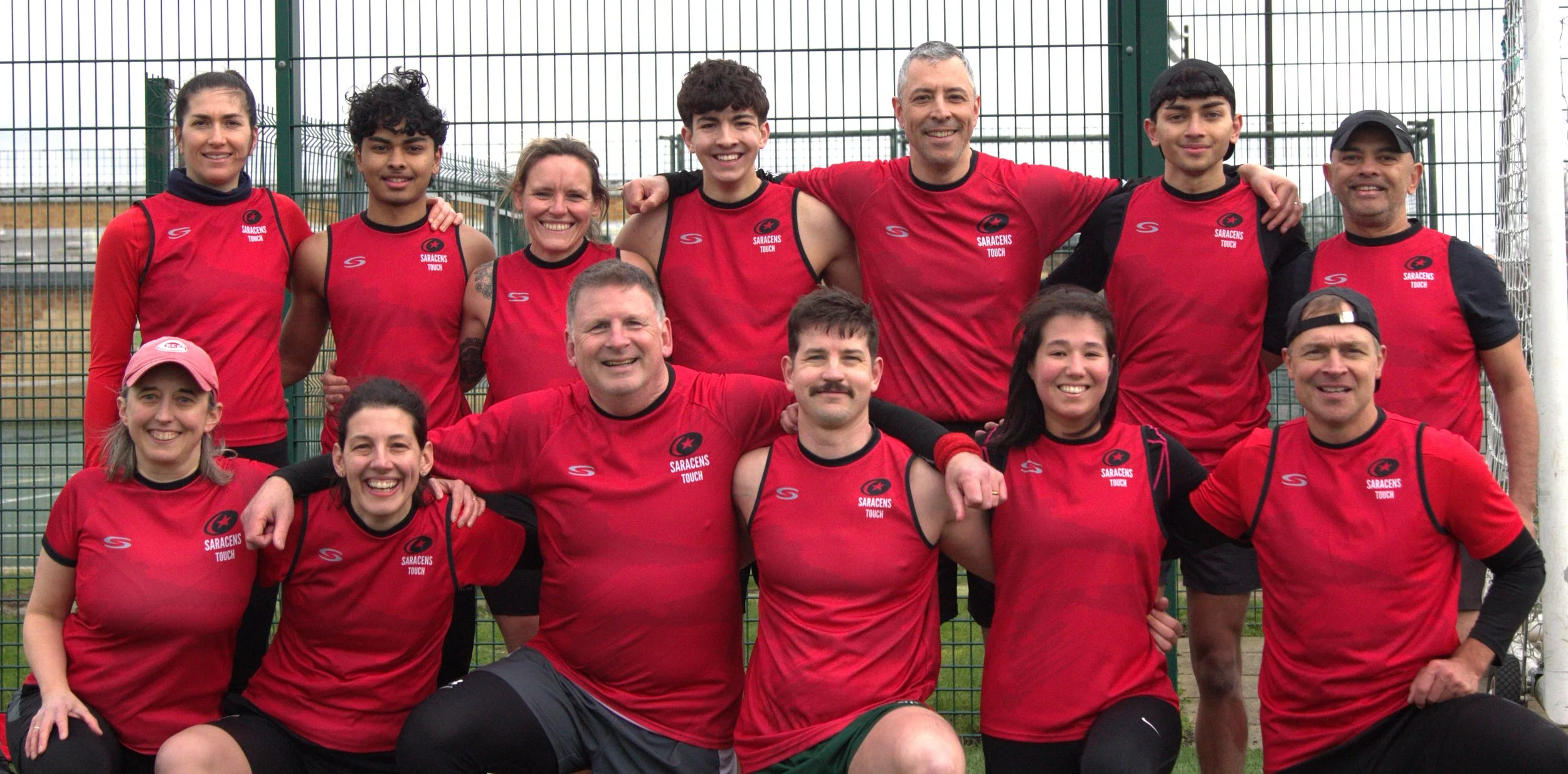 A group of eleven athletes and coaches wearing red sports jerseys, posing together in front of a fenced sports field, smiling for the camera.