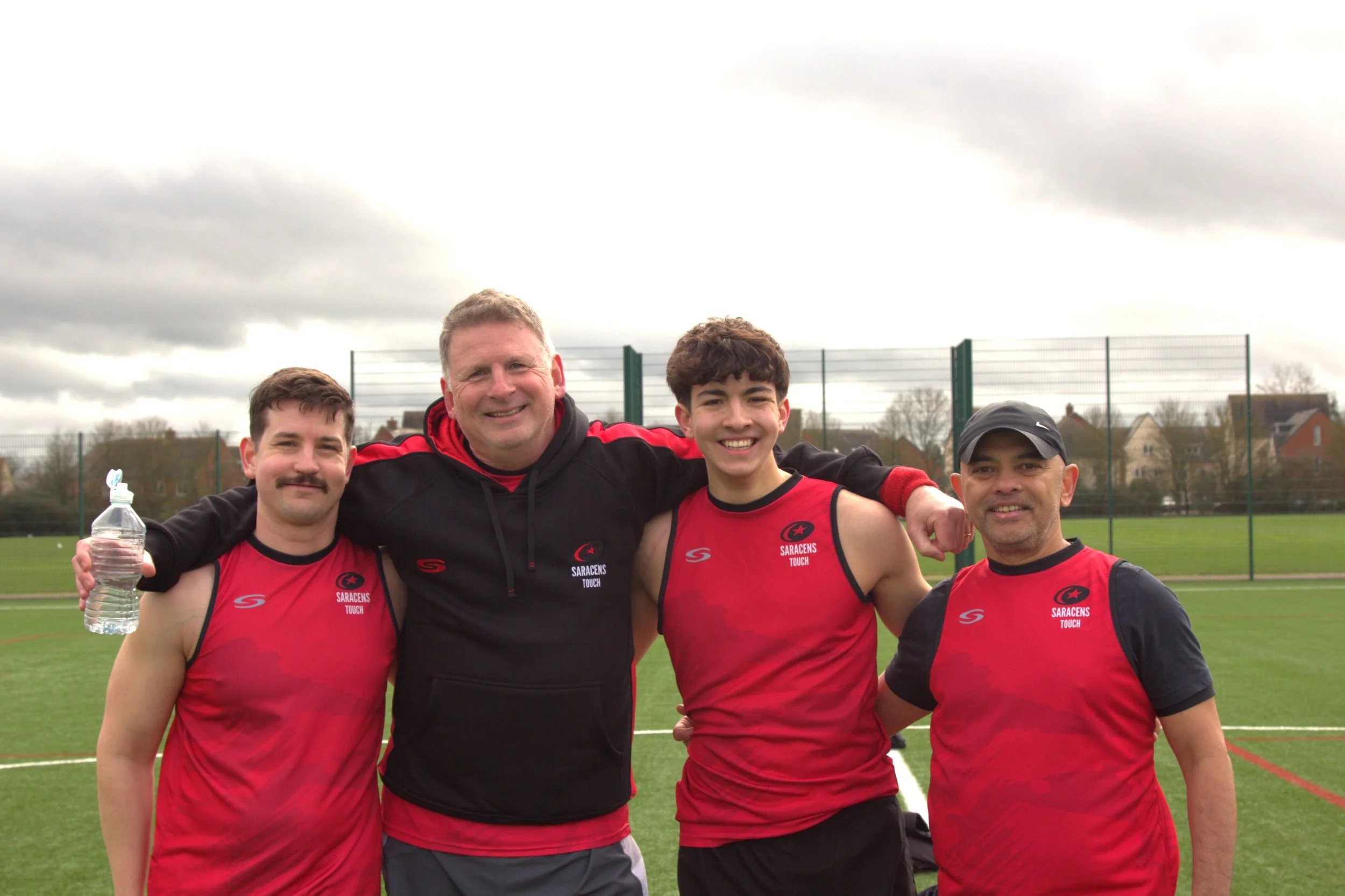 Four men standing together on a sports field, smiling, wearing red and black athletic uniforms with 'Sarayens Touch' logos, one holding a water bottle, others have their arms around each other's shoulders, with cloudy skies overhead.