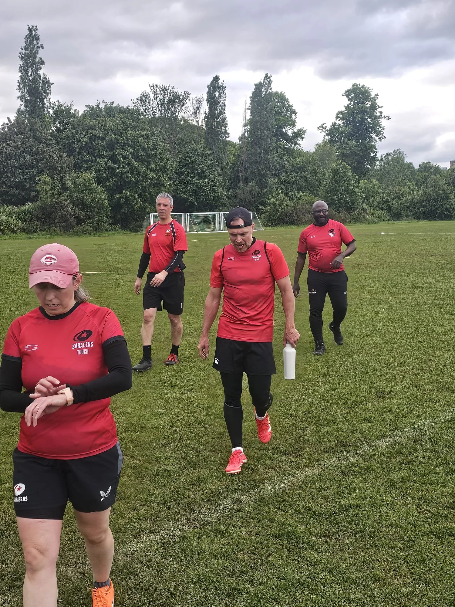 Group of people in red sports clothing walking on a grassy field with trees and overcast sky in the background.