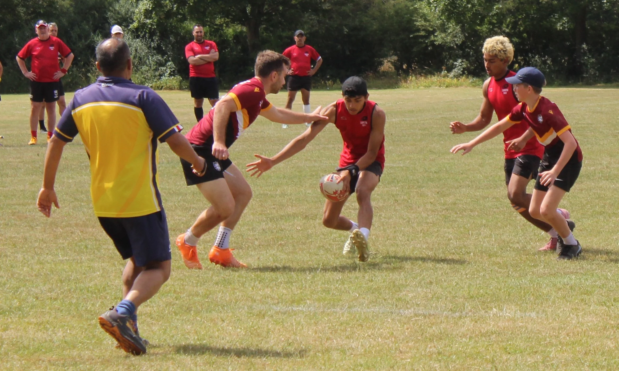 Young boys playing rugby with a ball on a grassy field, with adults watching in the background.