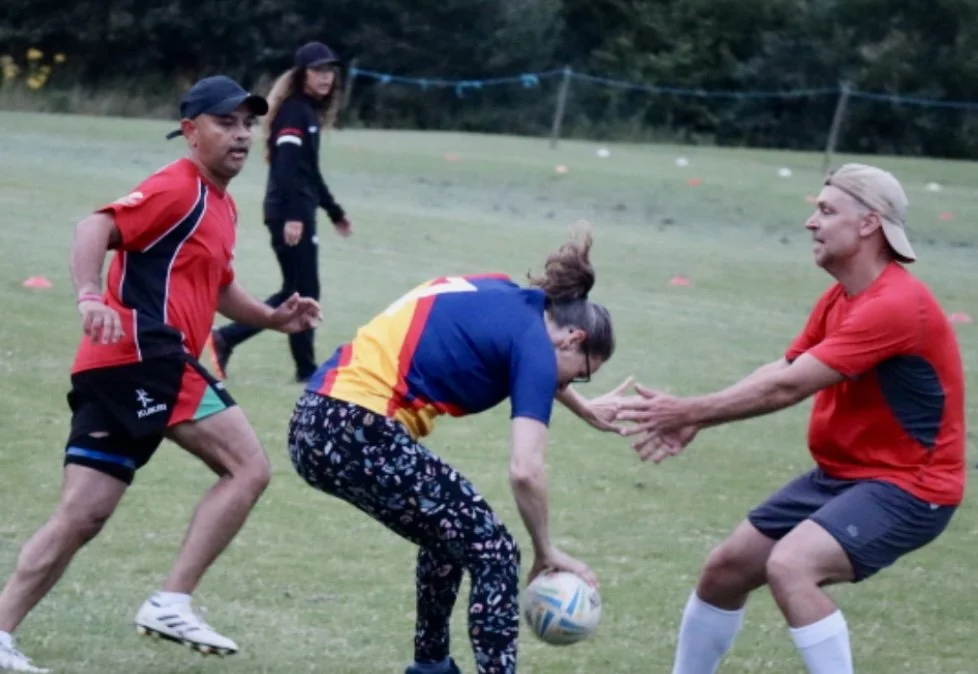 Four people playing rugby on a grassy field, with one woman passing the ball and three men engaging in the game.