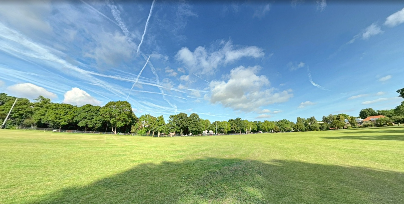 Open grassy field under a blue sky with scattered clouds and jet contrails, bordered by trees and distant houses.