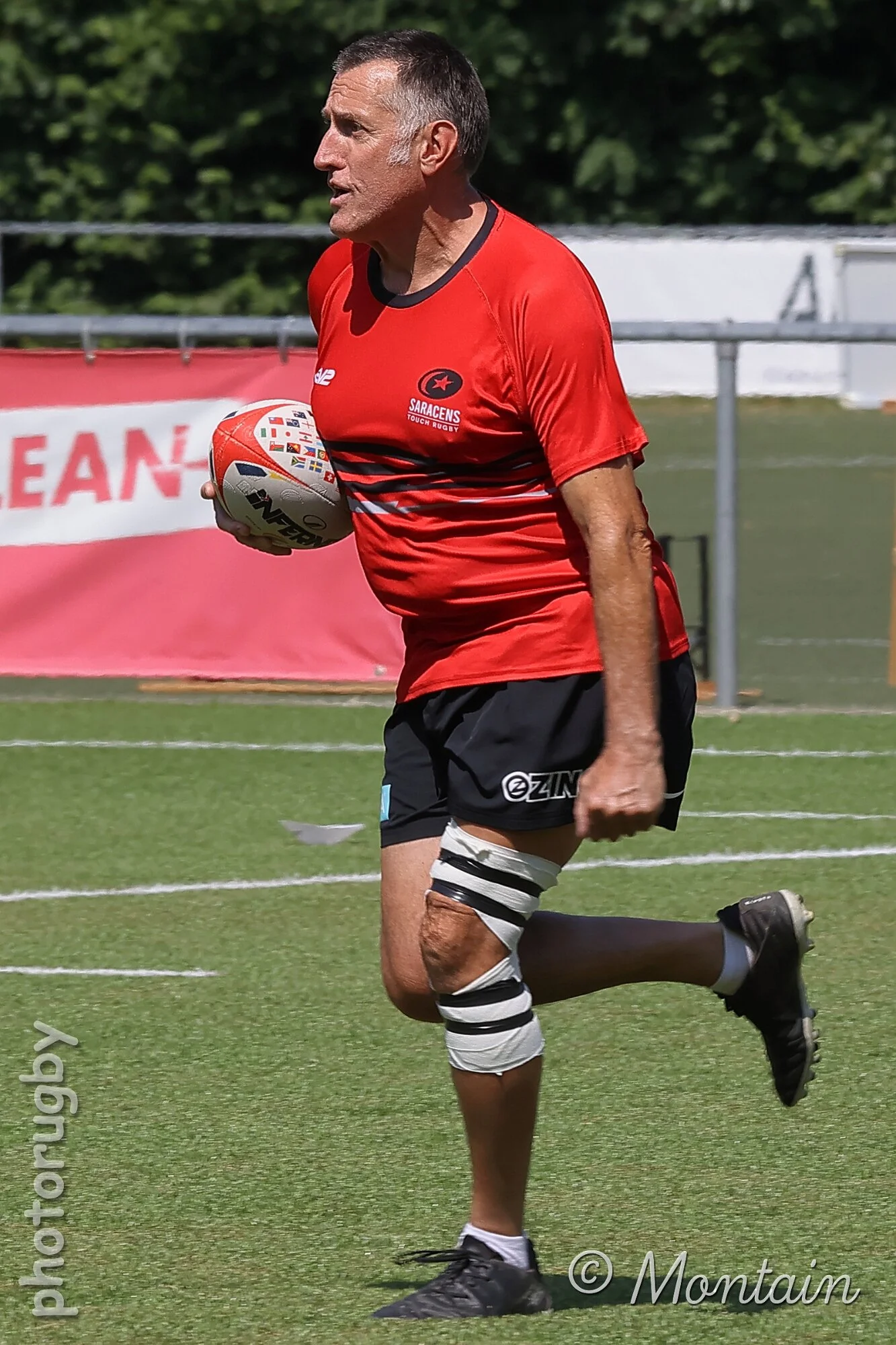 A man running on a sports field holding a rugby ball, wearing a red shirt with the Saracens logo, black shorts, and a knee brace.
