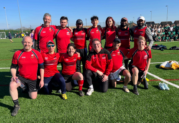 Group of eleven soccer players and coaches posing on a soccer field, wearing red and black uniforms, with a crowd in the background.