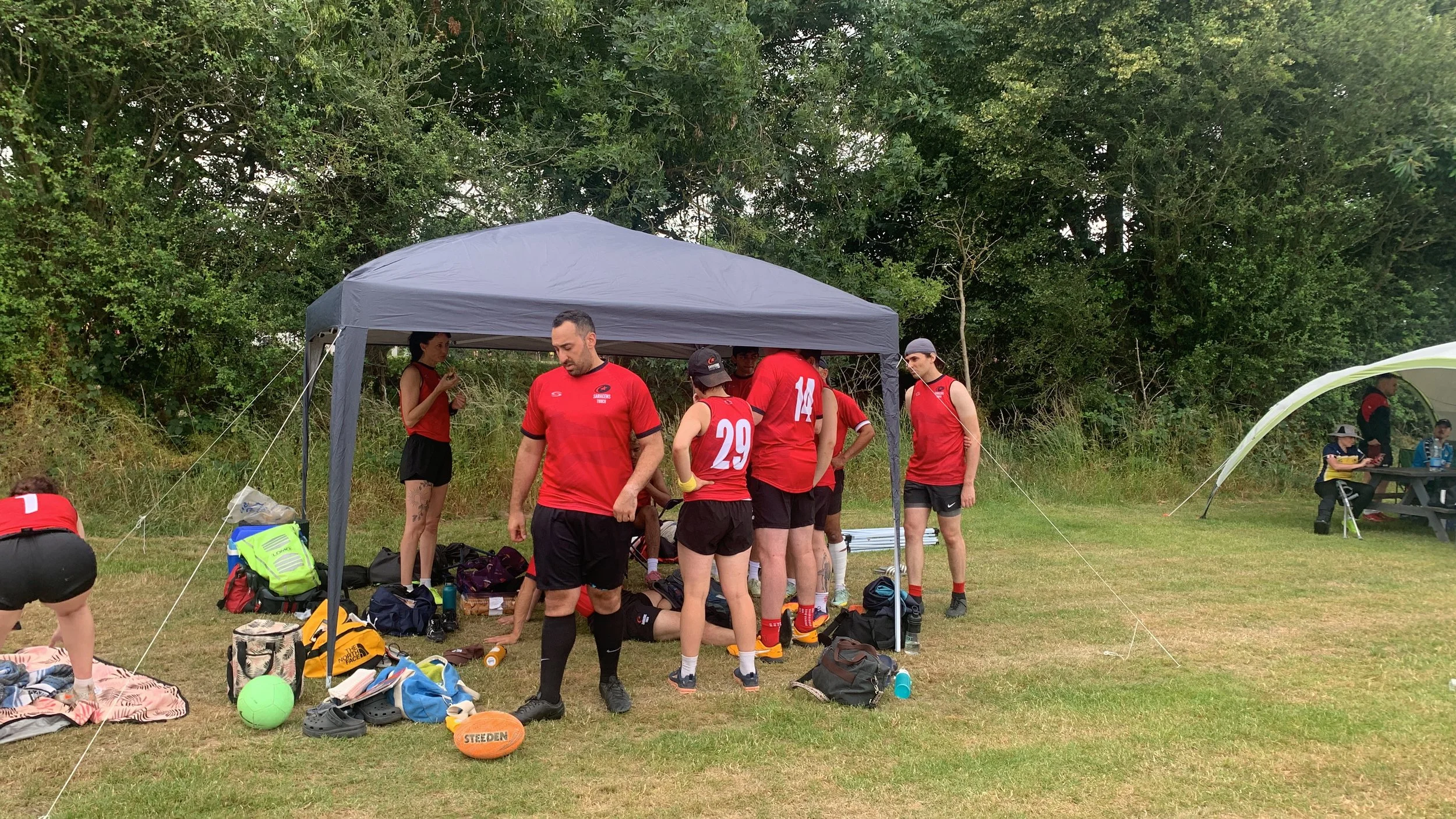 A group of athletes, dressed in red sportswear, are gathered under a gray canopy in a grassy outdoor area surrounded by trees. They are preparing or resting during a sporting event, with sports equipment and bags on the ground around them.