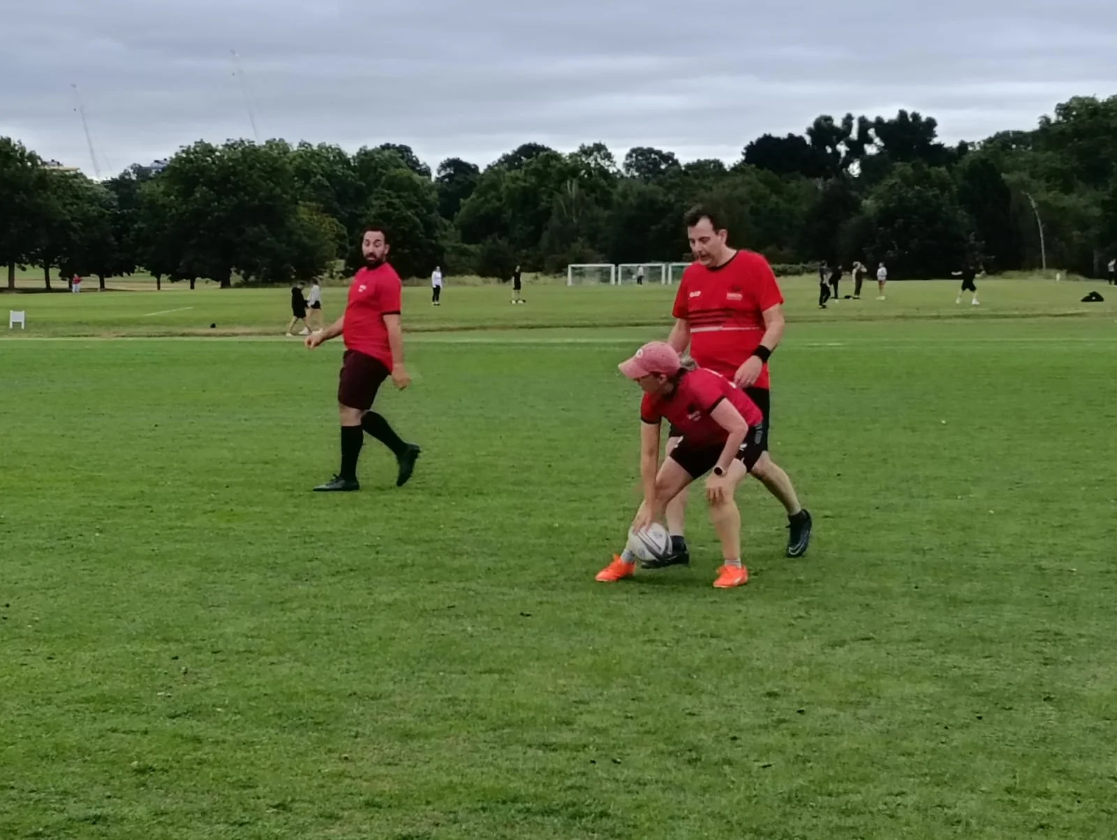 People playing rugby on a grassy field with trees in the background and cloudy sky overhead.