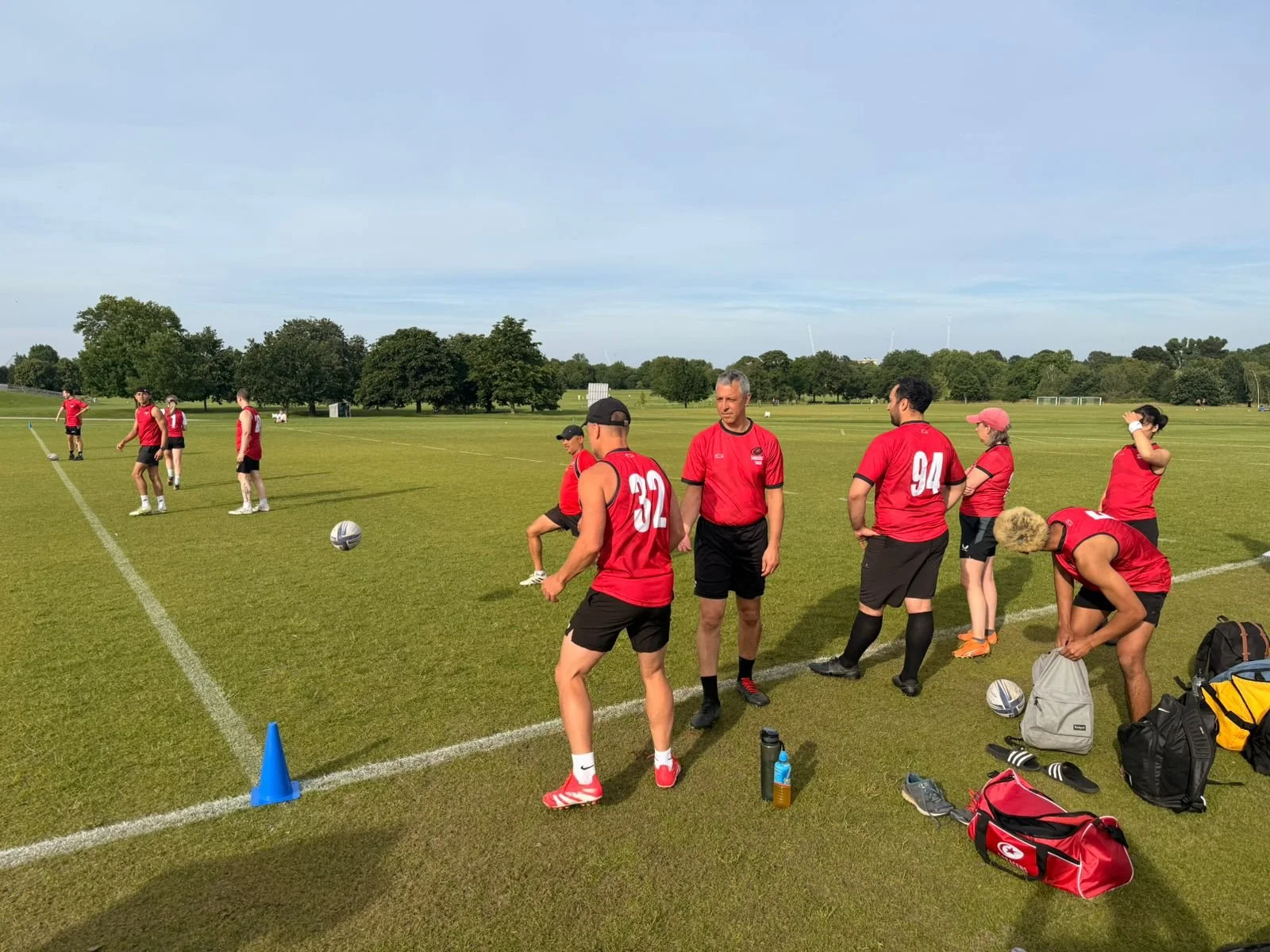 A group of rugby players in red jerseys and black shorts on a grassy field, some standing and some preparing for game or practice, with a few wearing hats and others adjusting equipment, during daytime with trees in the background.