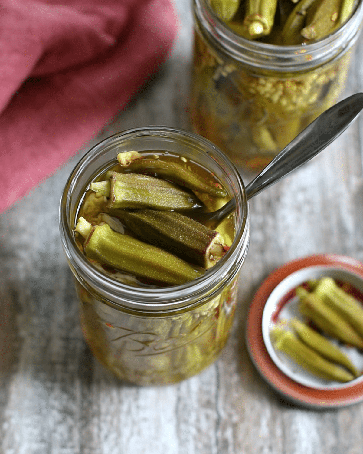 Close-up of a glass jar filled with pickled green okra, with a spoon inside, on a wooden surface with a pink cloth and other jars in the background.
