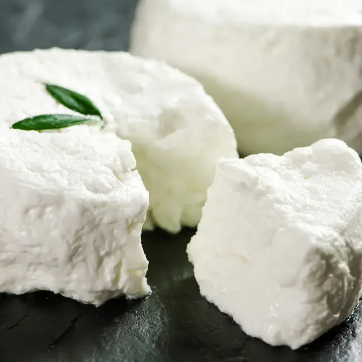 Close-up of a block of fresh white cheese with a small piece cut off, garnished with a small green leaf, on a dark surface.