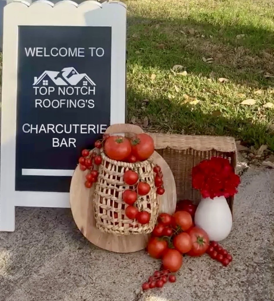 Sign welcoming visitors to Top Notch Roofing's charcuterie bar, with tomatoes and red berries arranged in baskets and vases outside.