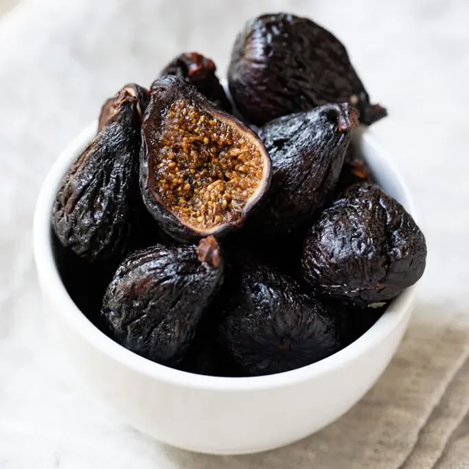A white bowl filled with dark purple or black figs, one of which is cut open showing its juicy, seeded interior.