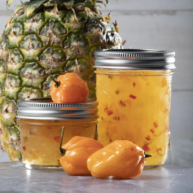 Pineapple and jars of pineapple jam with fresh habanero peppers on a gray surface.