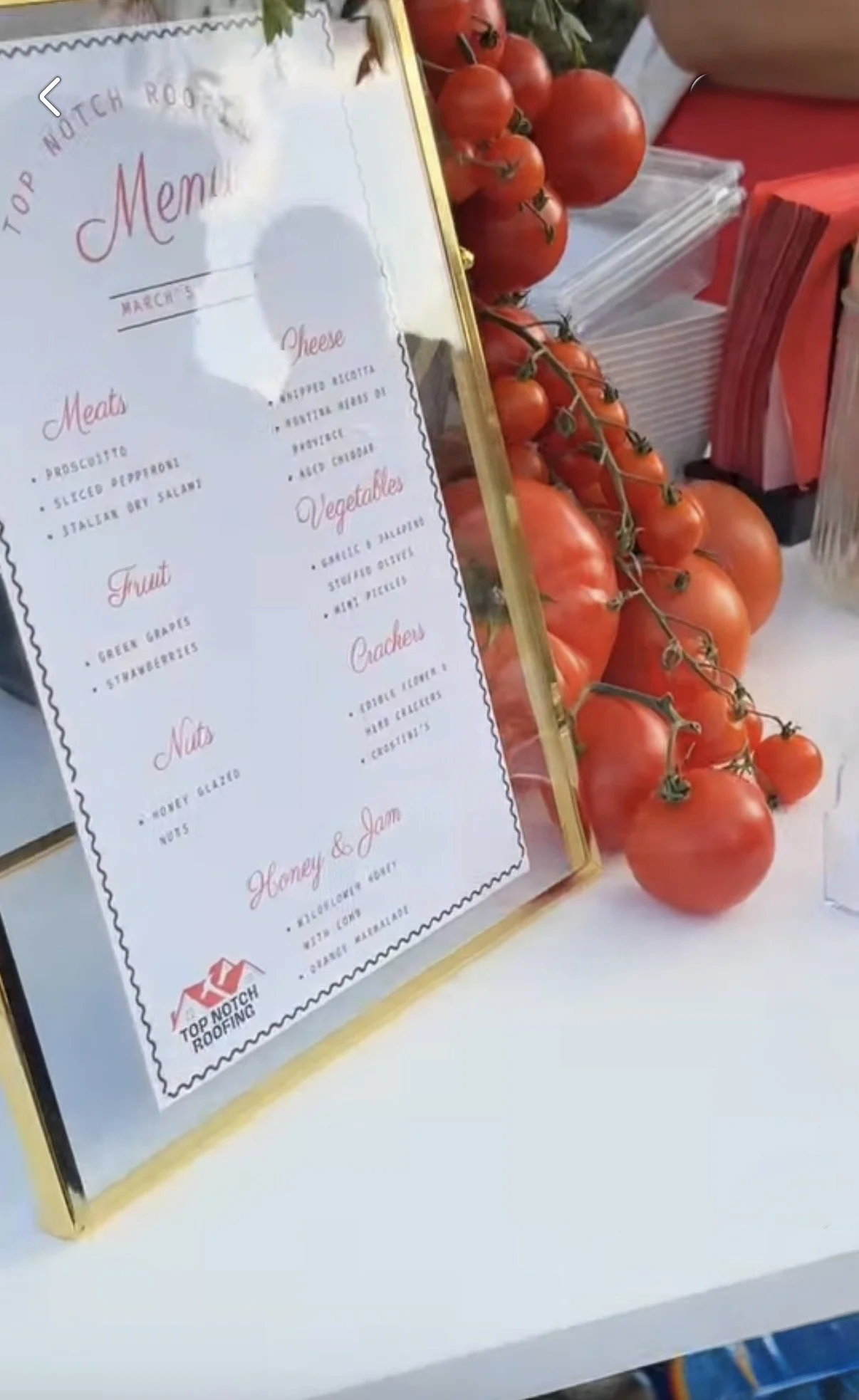 A menu board with red text and various food categories, placed beside a bunch of fresh red tomatoes on a white table, at what appears to be a food stand or market.
