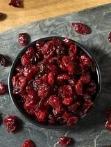 A small black bowl filled with dried, wrinkled cranberries on a dark slate surface, with some around the bowl.
