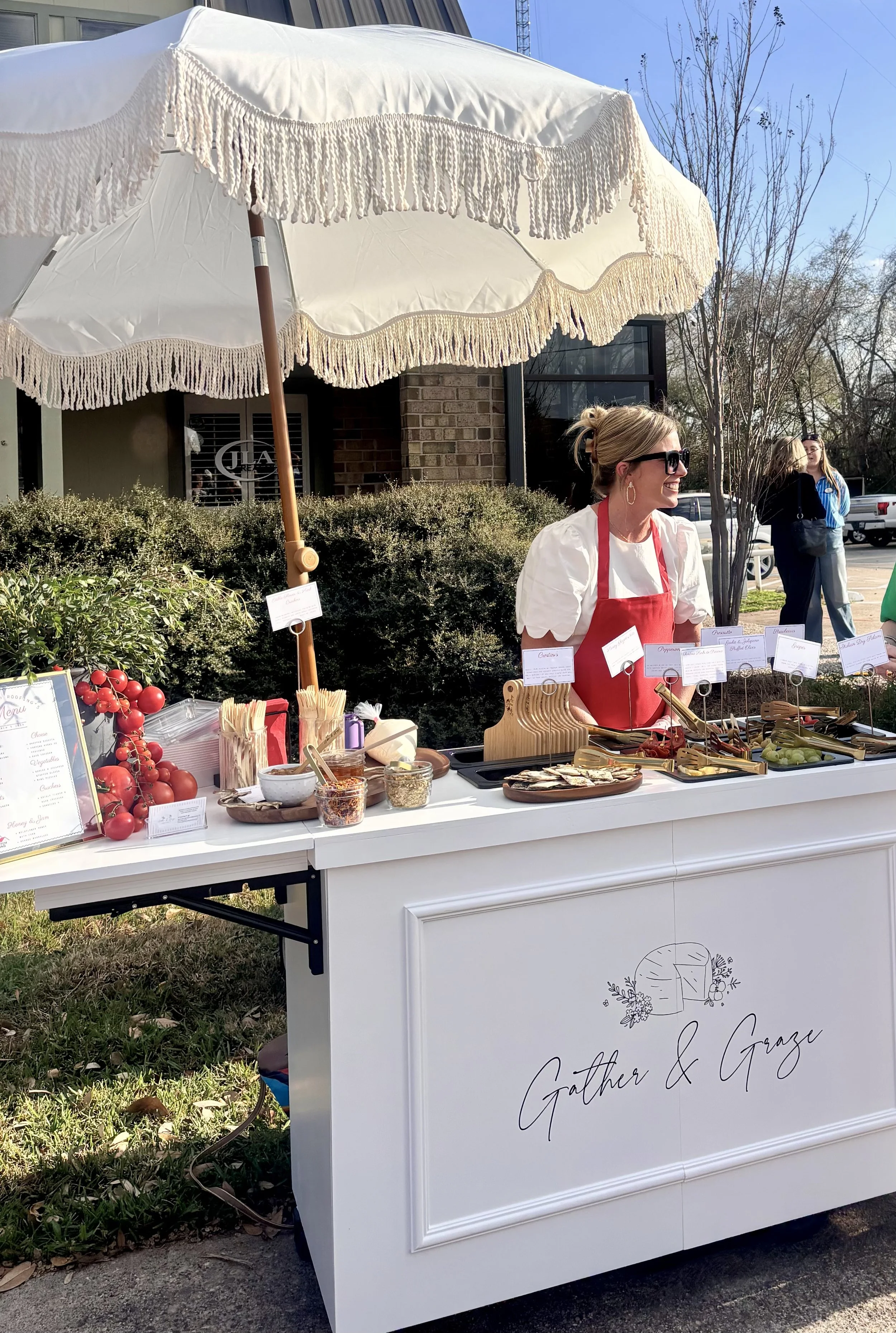 A woman wearing a white top and red apron standing at a white outdoor food stand with a large white fringed umbrella. The stand has various food items, condiments, and small signs. The stand features the logo and text ‘Gather & Graze’.