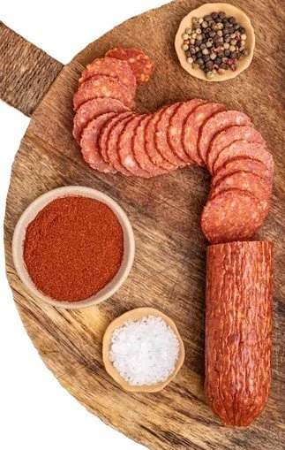 Sliced cured sausage, a whole sausage, ground spices in a bowl, and salt in a small bowl on a wooden cutting board.
