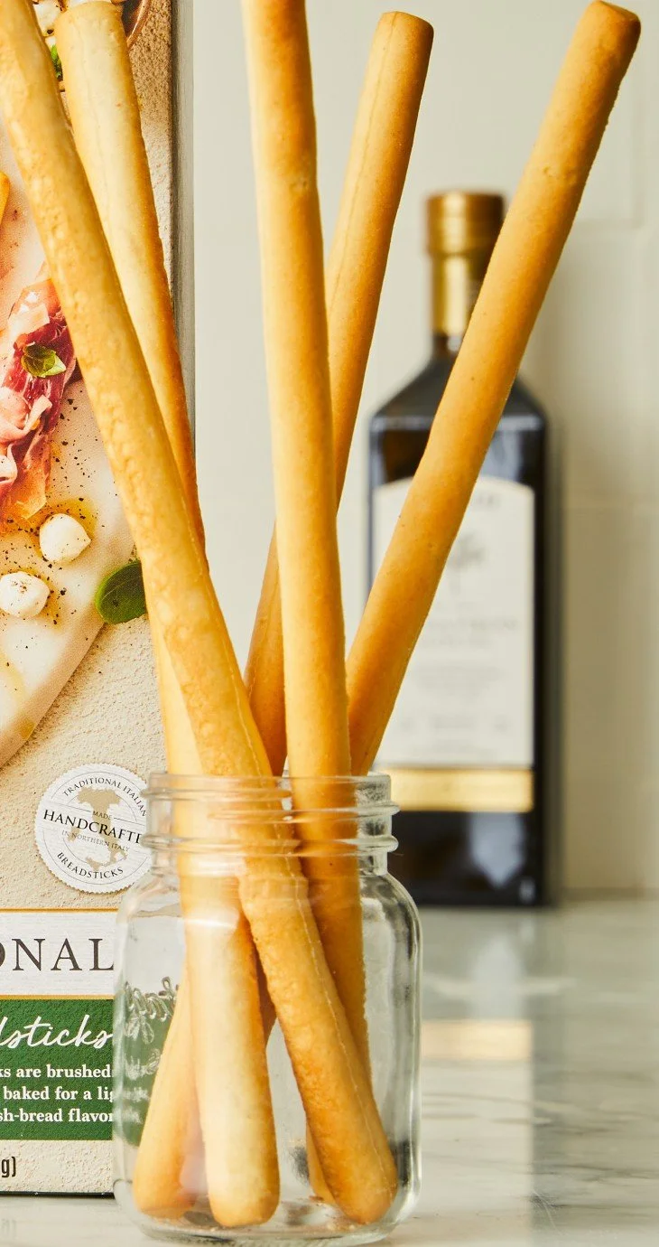 A glass jar holding several breadsticks on a kitchen countertop. In the background, there is a blurred dark bottle and part of a pizza box.