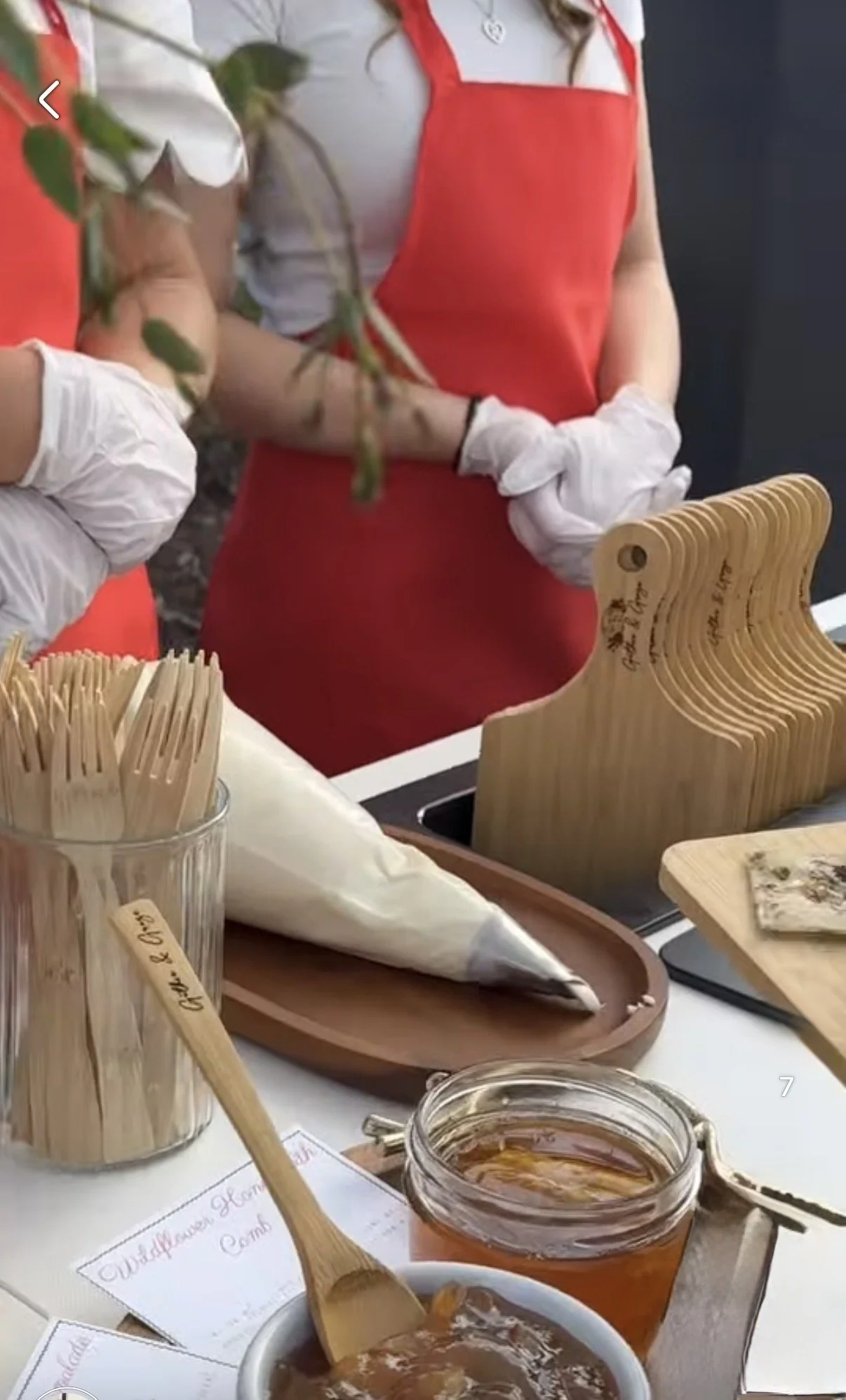 A person wearing a red apron and white gloves stands behind a counter with wooden utensils, a bag of flour, a jar of honey, and various baking tools, with a blurred plant in the foreground.