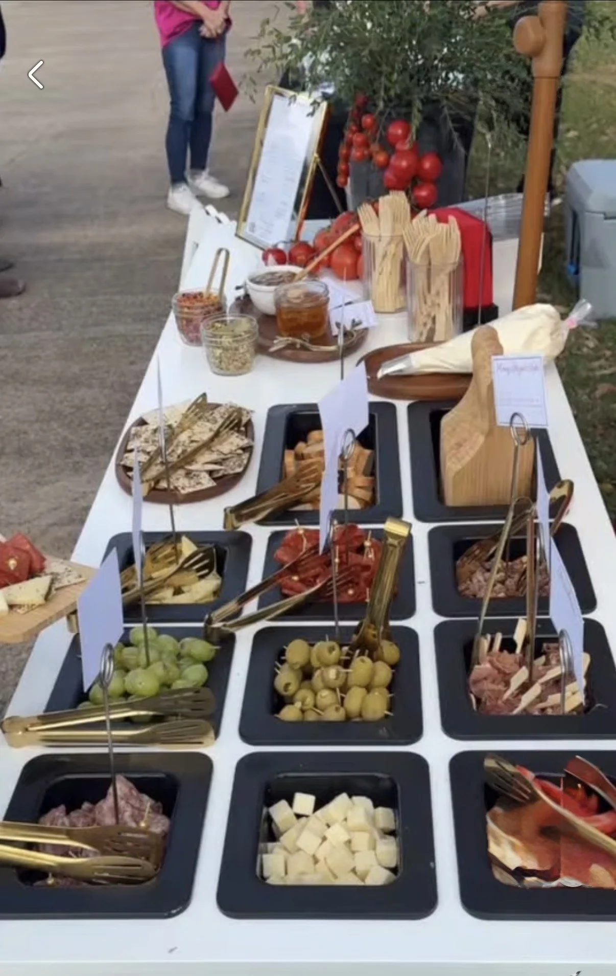 A salad bar with various bowls of cheeses, olives, sliced meats, and toppings, with tongs and labels for each item.