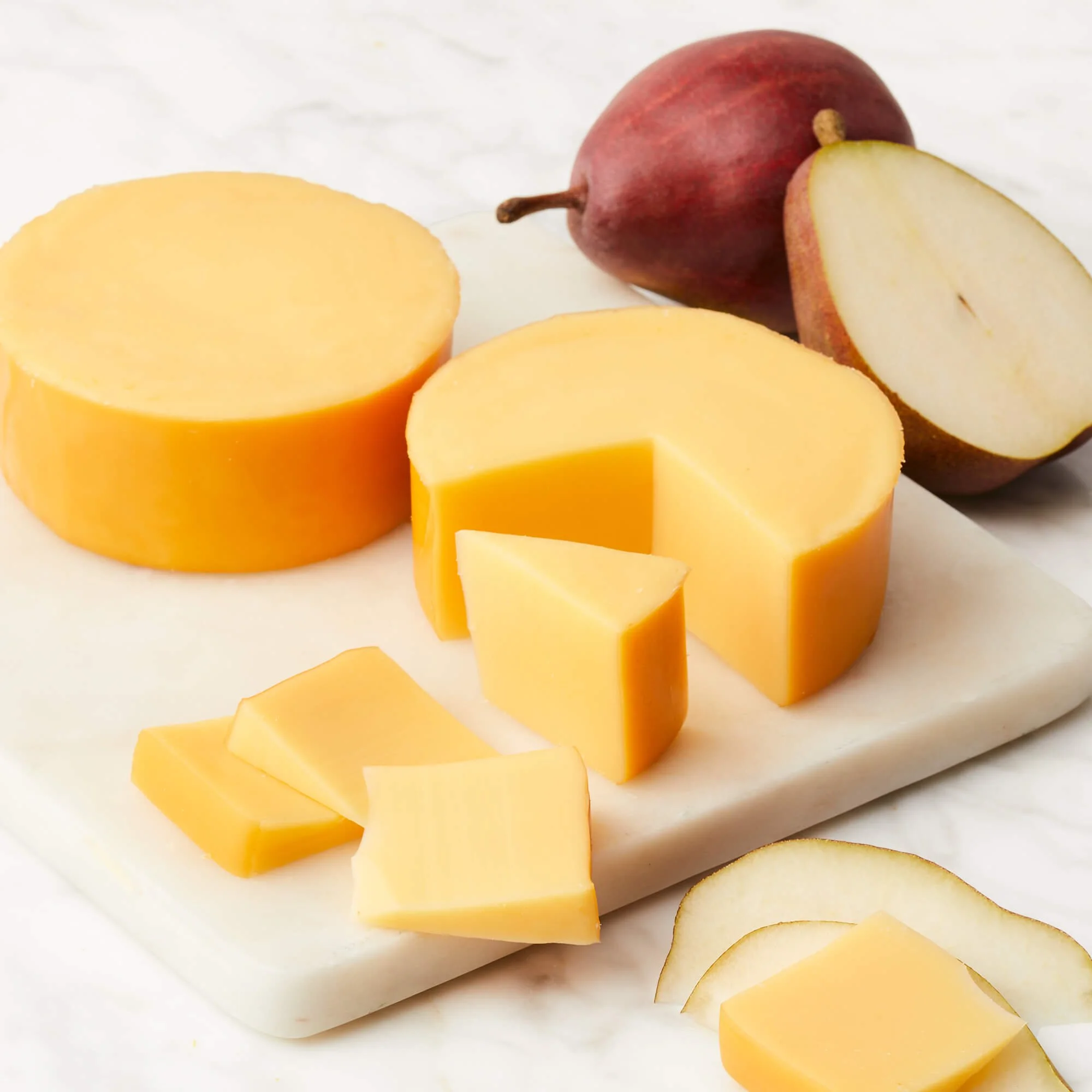 Slices of yellow cheese on a white marble surface with a red pear, halved, in the background.