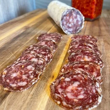 Sliced cured sausage on a wooden cutting board, with a whole sausage in the background.