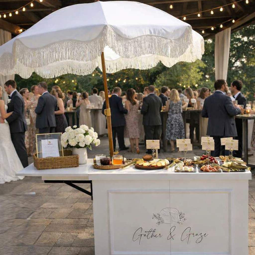 A wedding reception outdoor setting with guests socializing in the background. In the foreground, a white table with a large white parasol featuring fringe, a basket of flowers, and a tray of snacks and drinks.